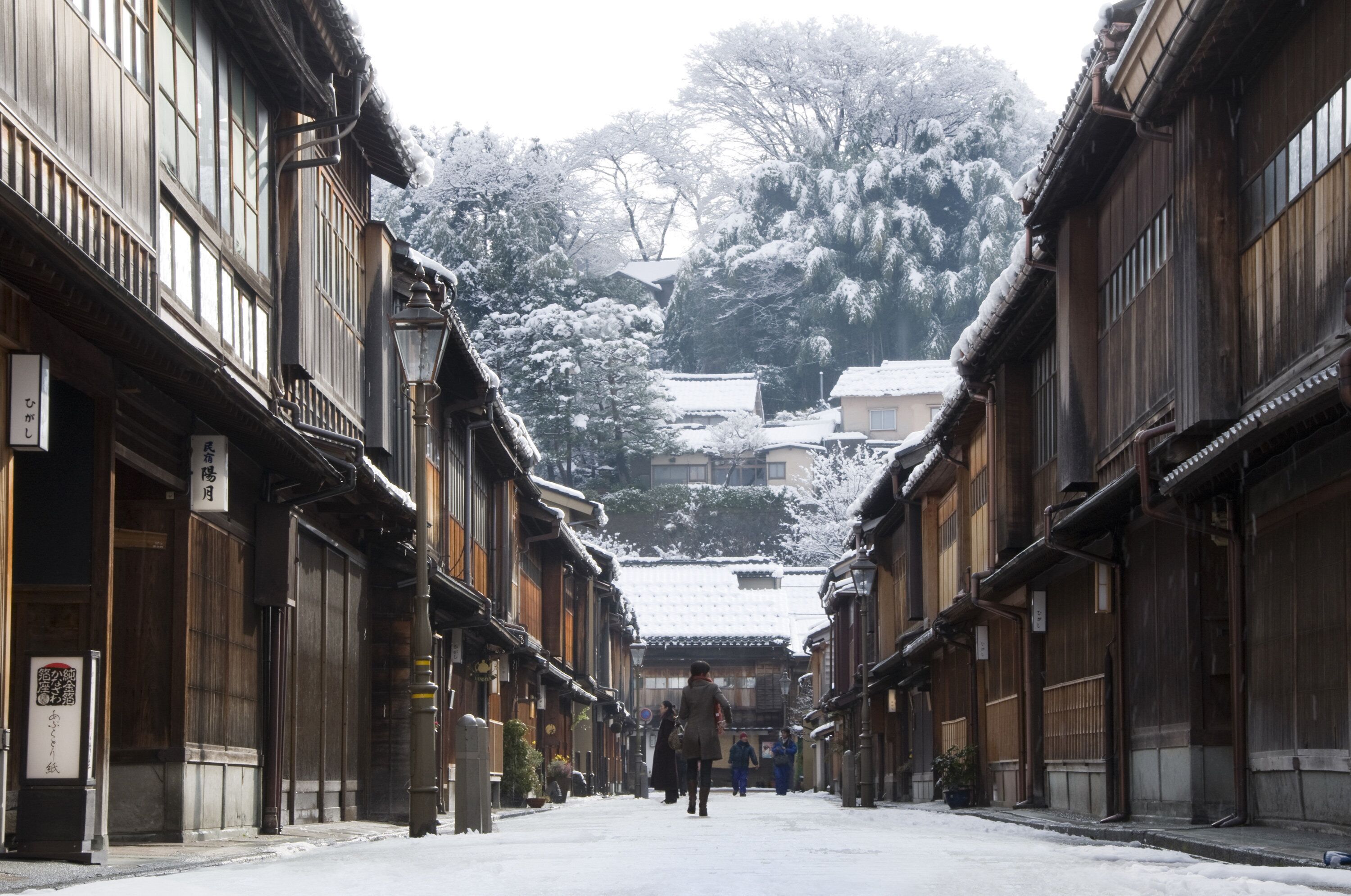 A traditional wooden street with buildings on either side, a person walking down the street, and snow-covered trees in the background.