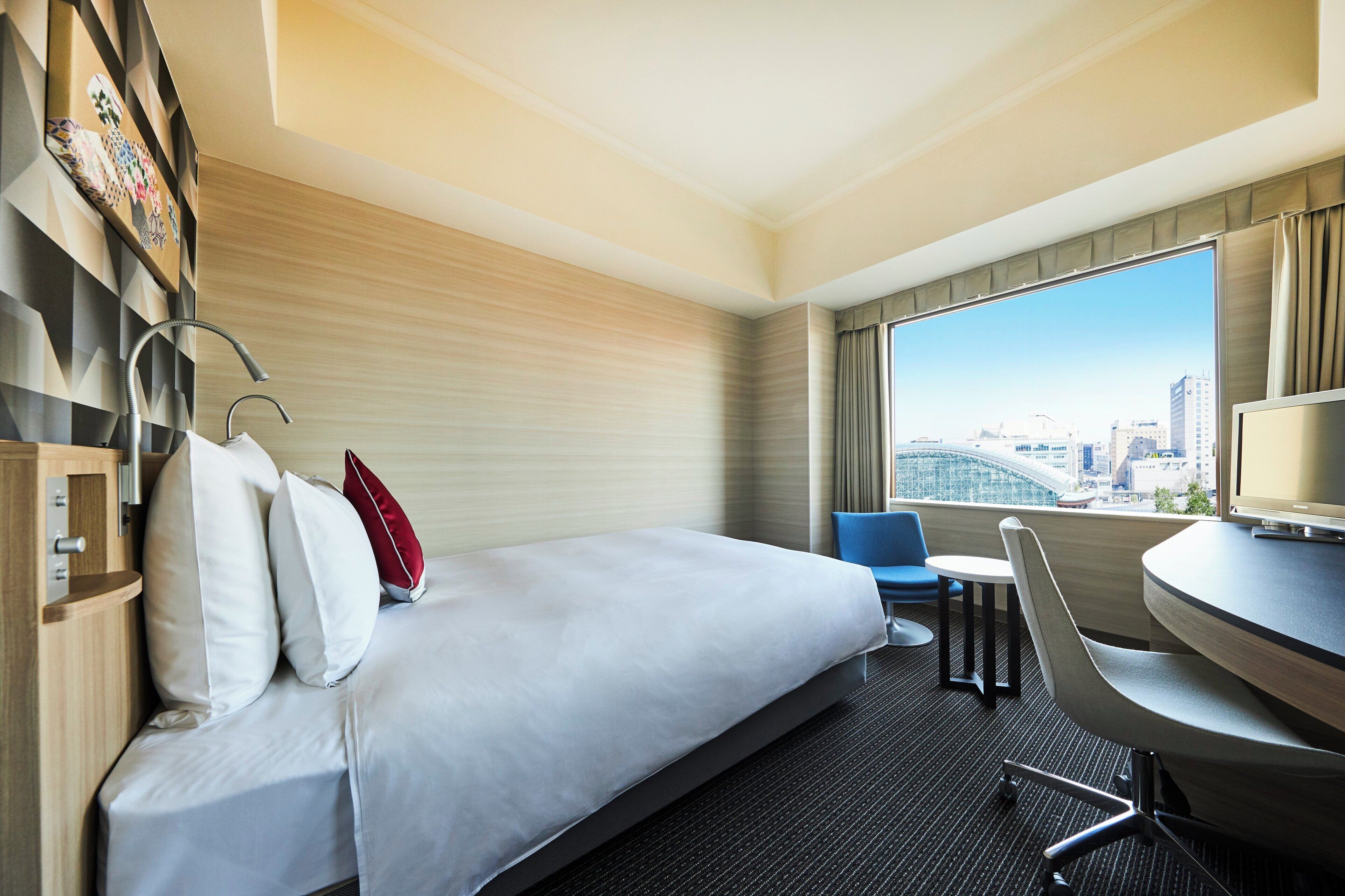 A hotel room with a bed, a desk, a chair, and a large window with a city view.