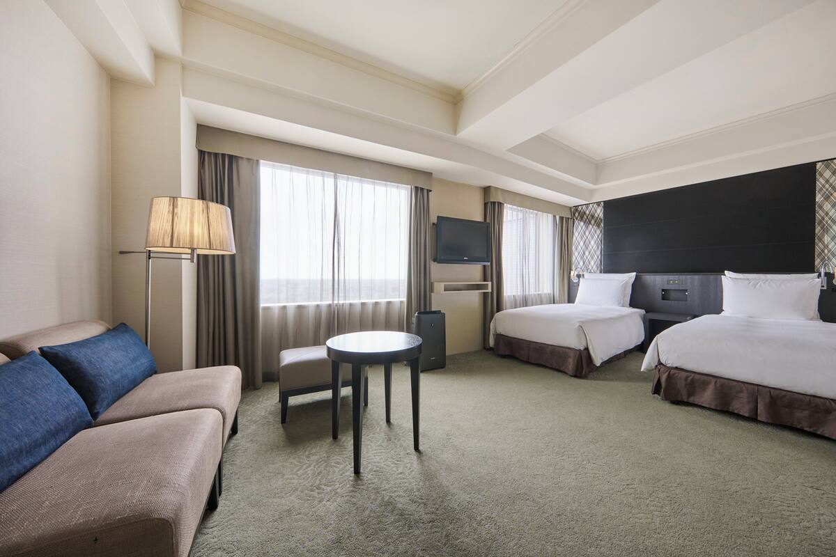 Une chambre d’hôtel avec un canapé, deux lits, une télévision, une petite table et une fenêtre avec des rideaux.