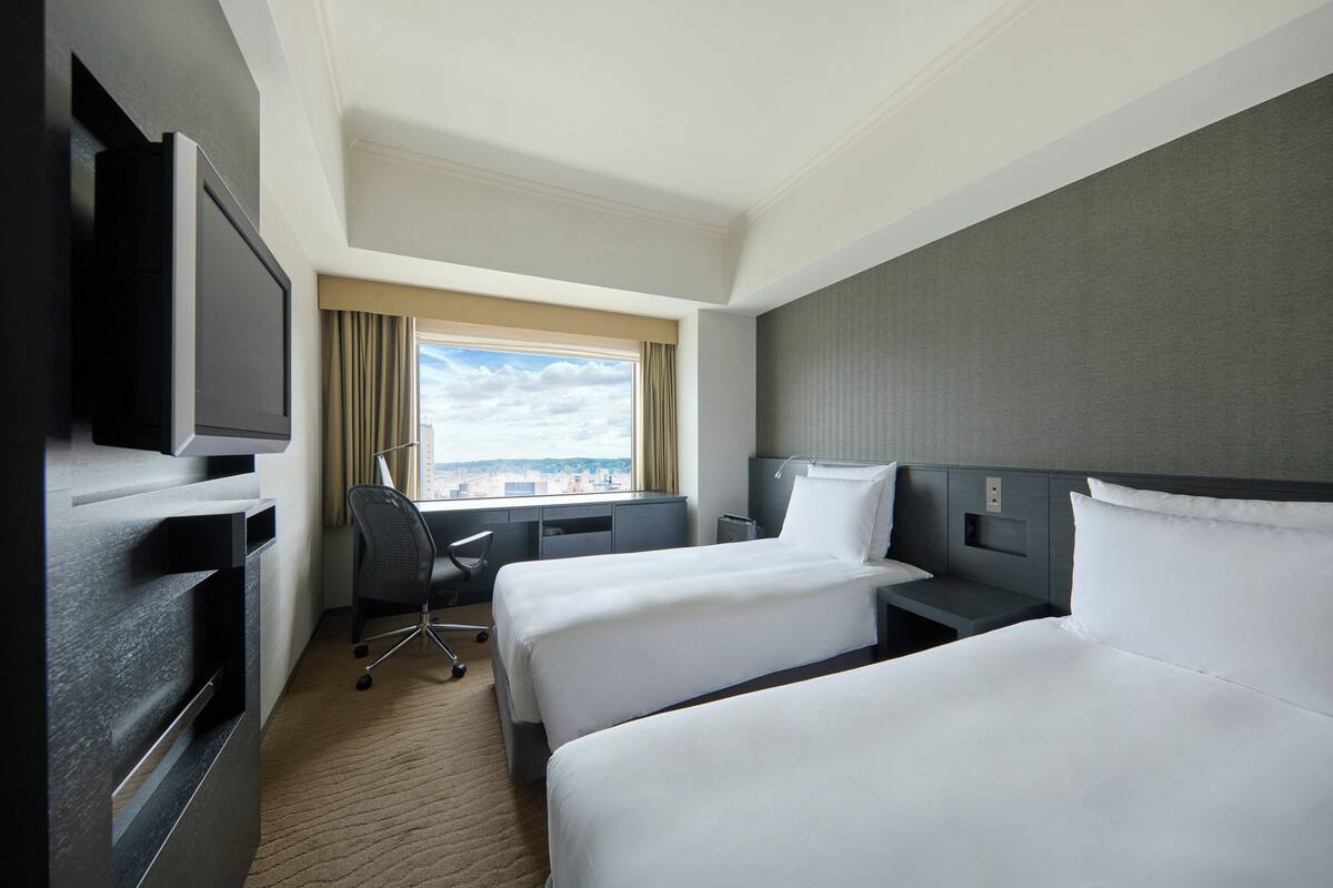 Une chambre d’hôtel avec deux lits, un bureau, une chaise, une télévision et une fenêtre donnant sur la ville.