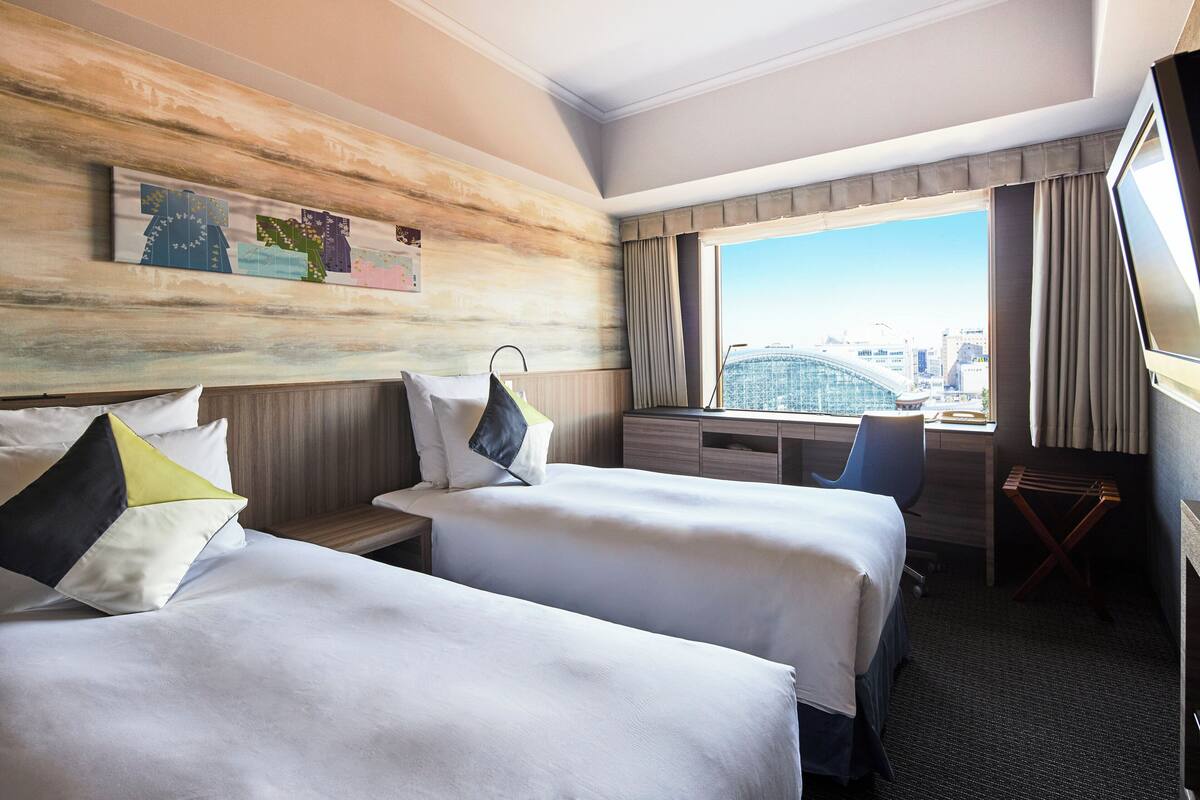 Une chambre d’hôtel avec deux lits, un bureau, une chaise et une grande fenêtre donnant sur la ville.