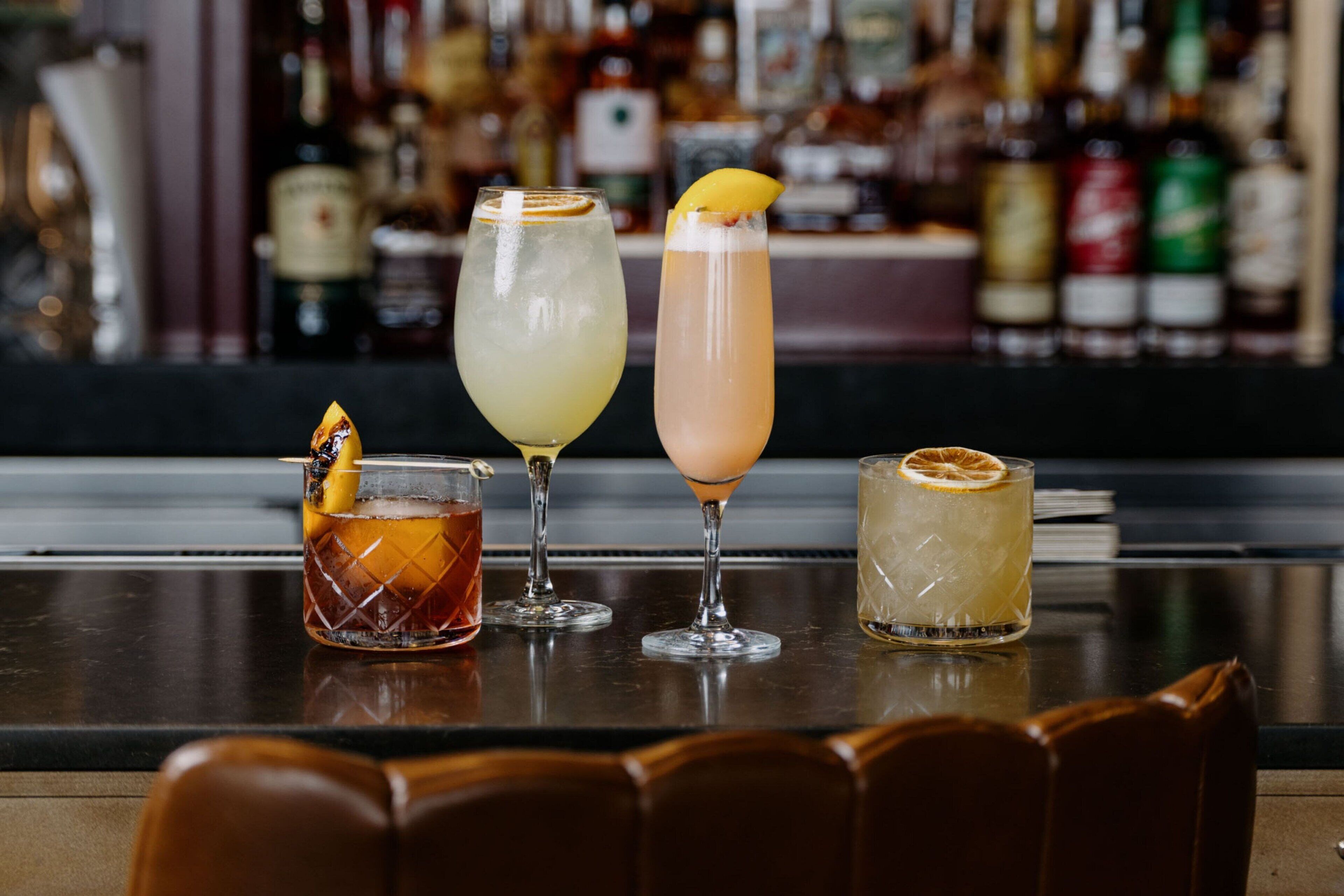 A bar with four different cocktails on a counter, with a blurred background of various bottles.