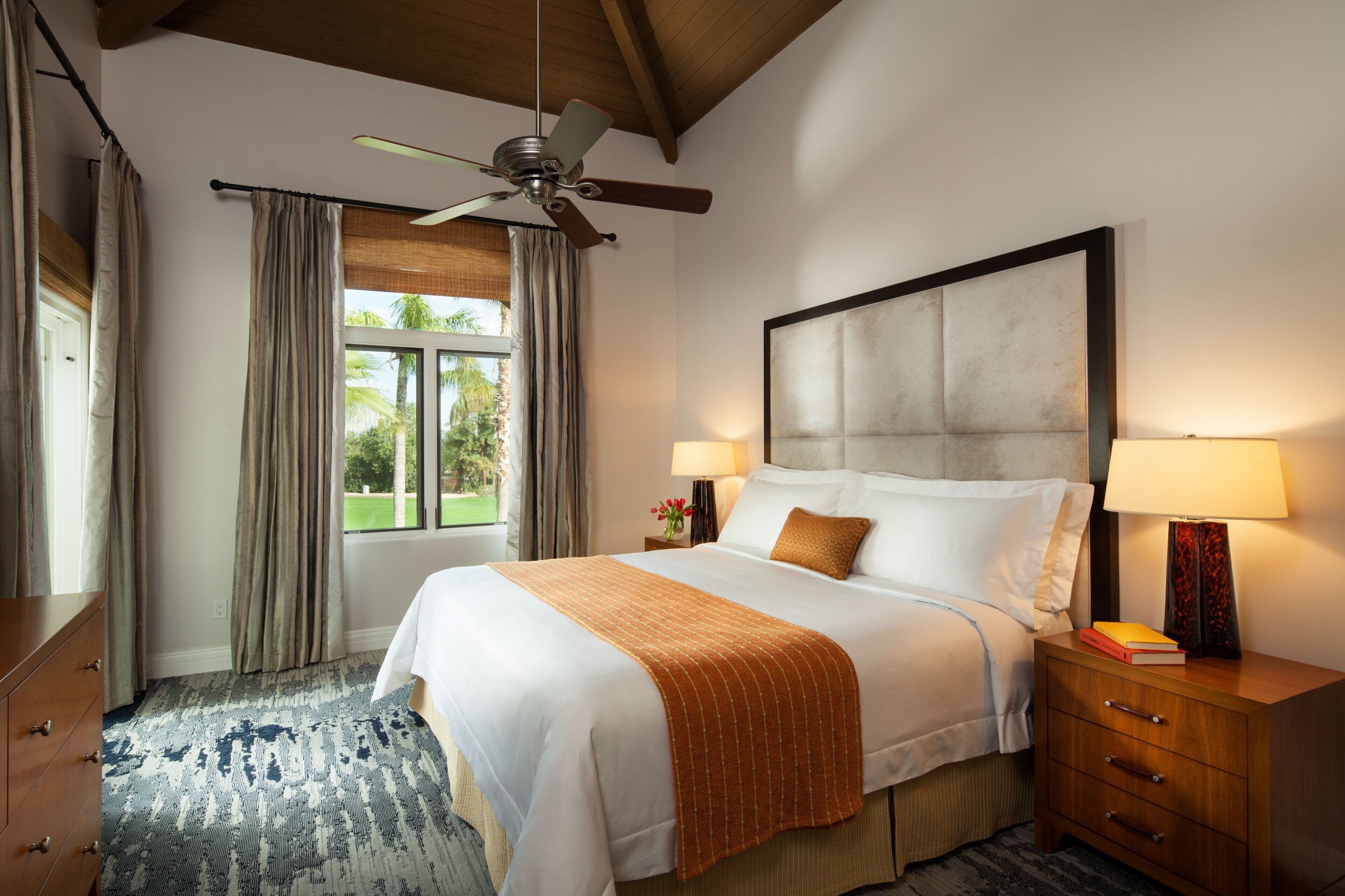 A hotel room with a bed, bedside lamps, a nightstand, a wooden dresser, a ceiling fan, and a window with curtains.