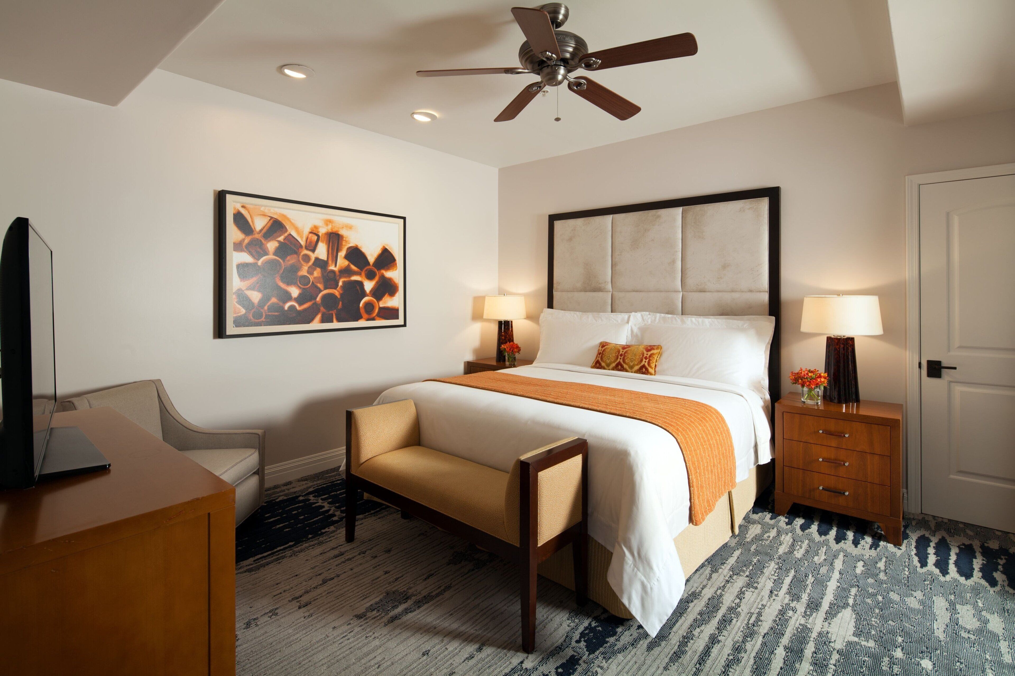 A hotel room with a bed, bedside tables, a desk, a chair, a ceiling fan, and a patterned rug.