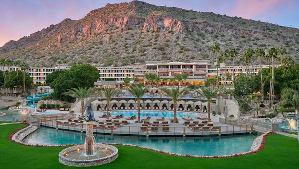 A resort with a large swimming pool, palm trees, and a mountainous backdrop.