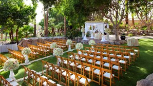 A wedding ceremony setup with rows of wooden chairs and white tablecloths, surrounded by greenery and rocks.