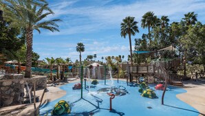 Un parc aquatique doté de nombreuses attractions aquatiques, notamment un toboggan et une aire de jeux avec jets d’eau, le tout entouré de palmiers et surplombé par un ciel bleu azur.