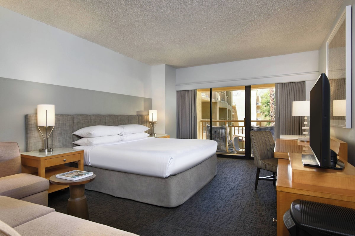 A hotel room with a large bed, a desk with a computer, a chair, a small table with a magazine, and a view of the outside balcony.