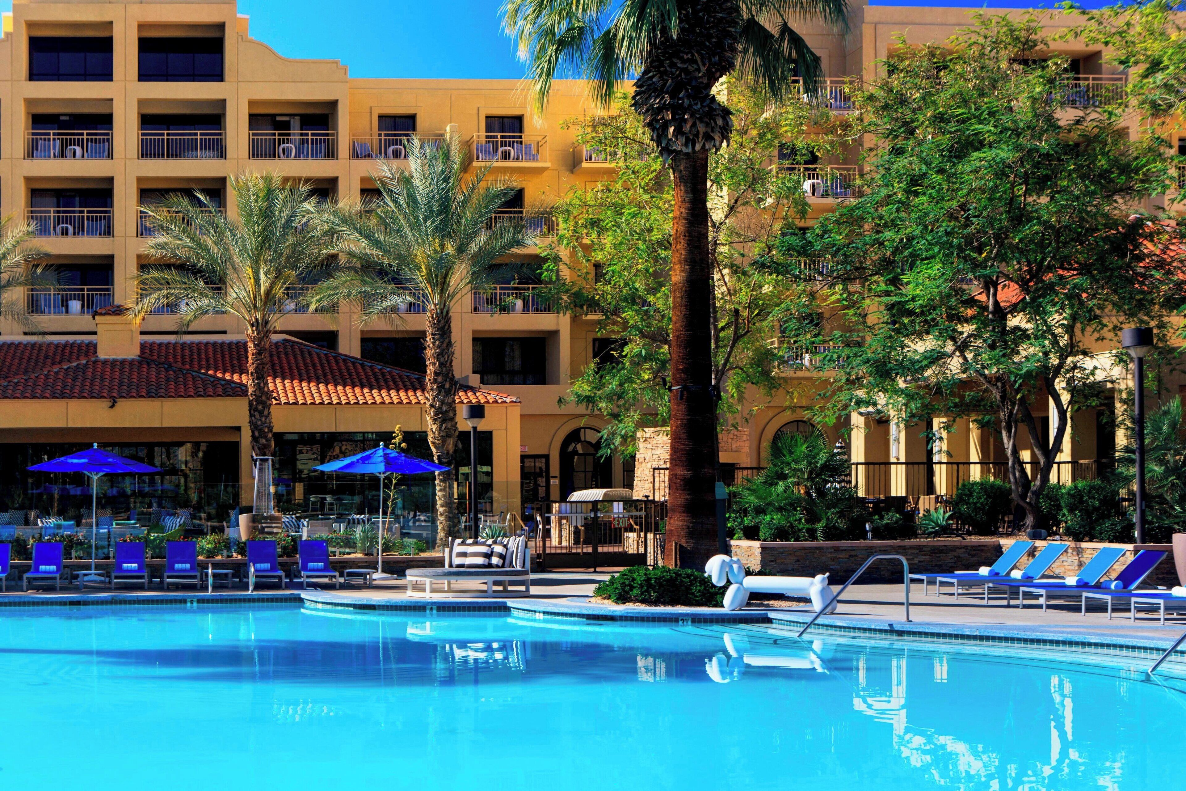 A hotel pool area with lounge chairs and umbrellas, surrounded by palm trees and a multi-story building with balconies.