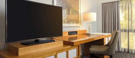 A hotel room with a flat-screen TV, a desk with a lamp, a chair, and a window with curtains.