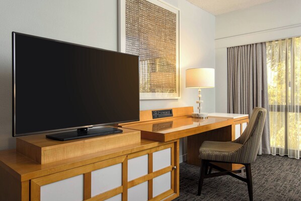 Habitación de hotel con televisión de pantalla plana, escritorio con lámpara, silla y ventana con cortinas.