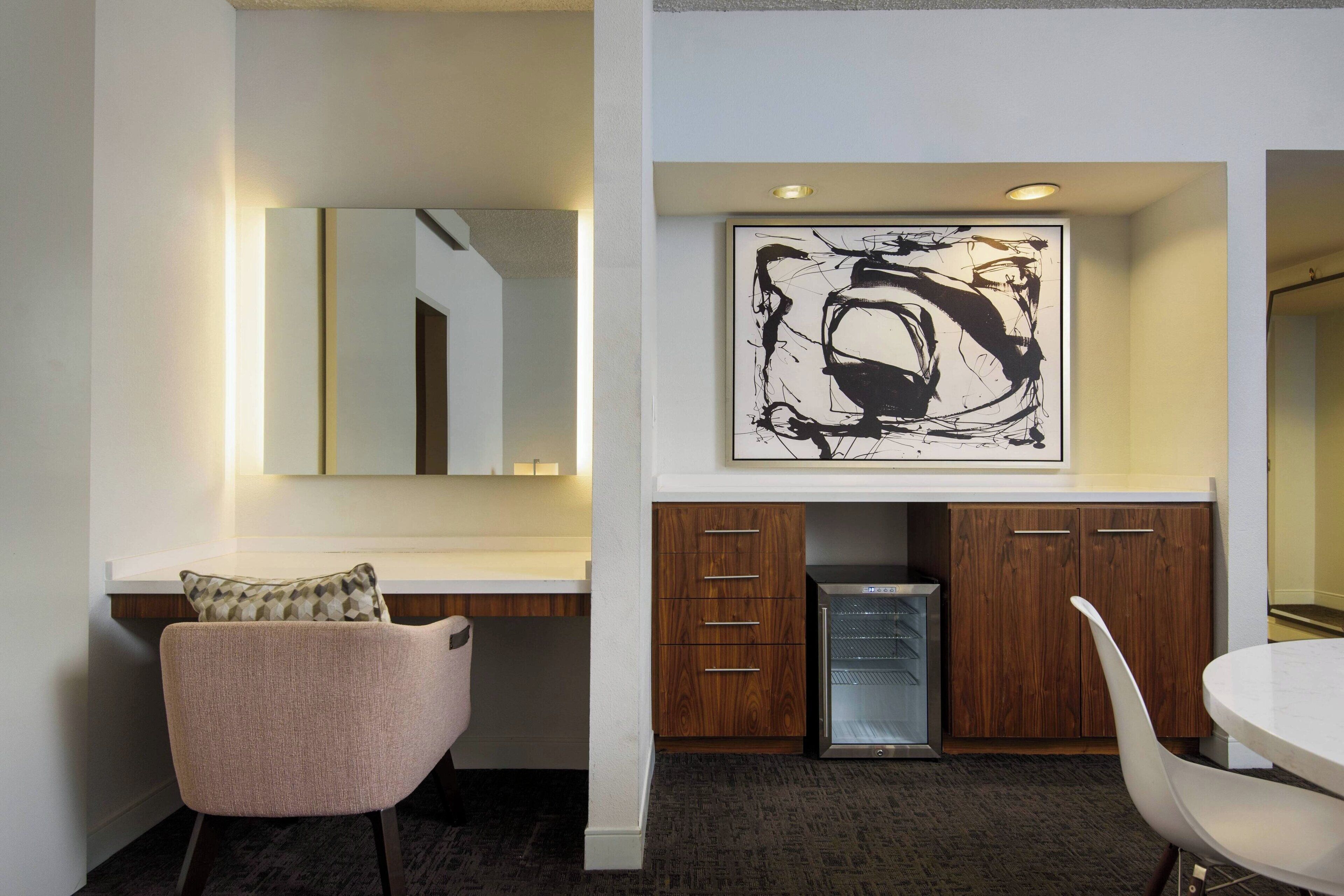 A modern hotel room with a large abstract painting, a wooden cabinet, a refrigerator, and a round dining table.