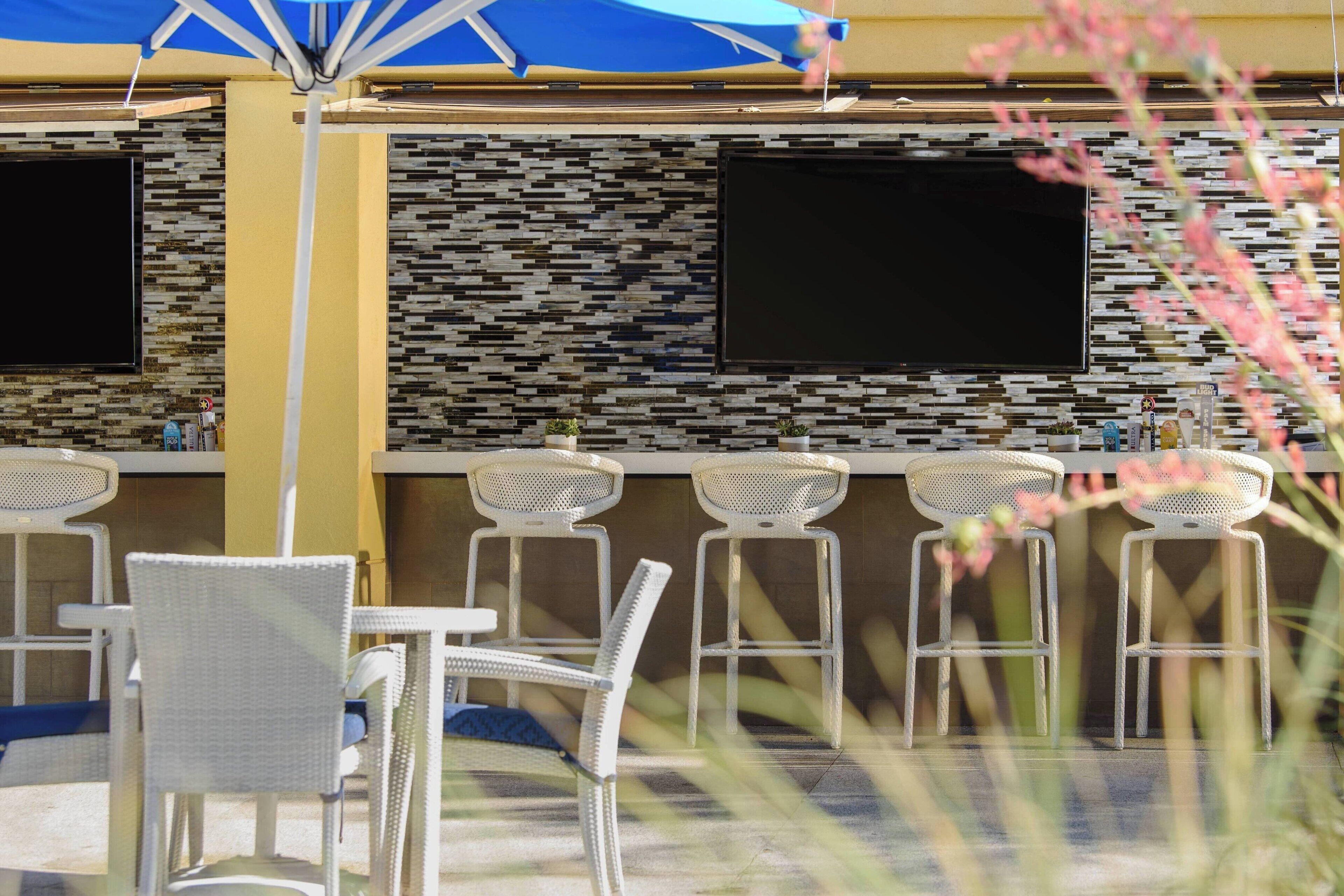 An outdoor bar area with white wicker chairs and a blue umbrella, featuring a tiled backsplash and two flat-screen TVs.