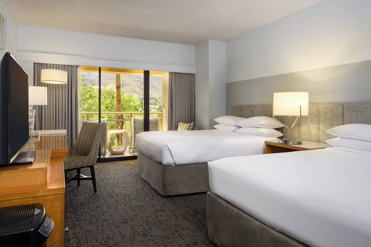 A hotel room with two beds, a desk with a TV, a chair, and a balcony with a view of greenery.