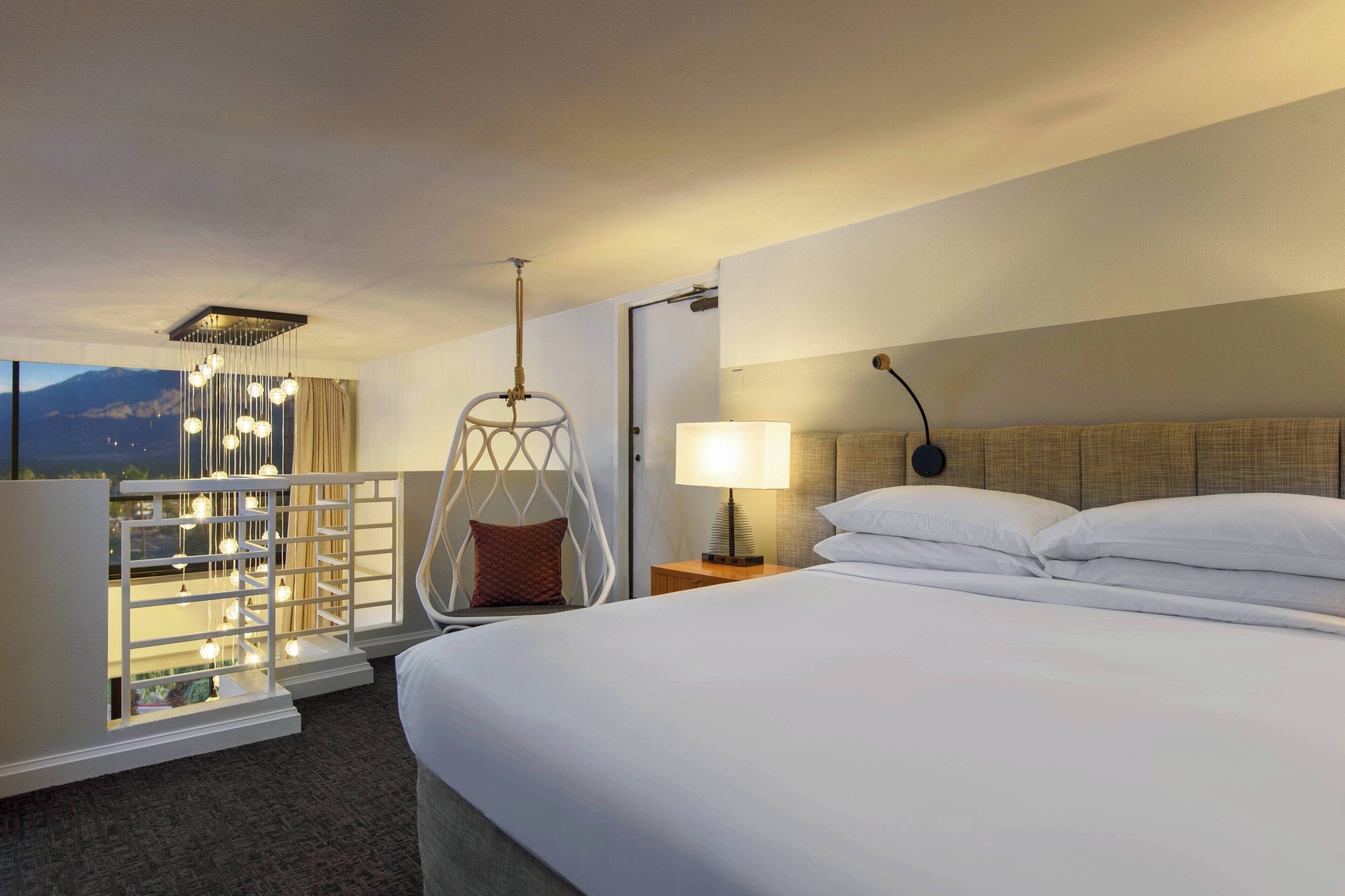 A hotel room with a large bed, a hanging chair, a bedside table with a lamp, and a view of the mountains.