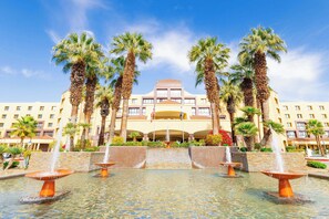 Un hotel con una fuente central, palmeras y un cielo azul despejado.