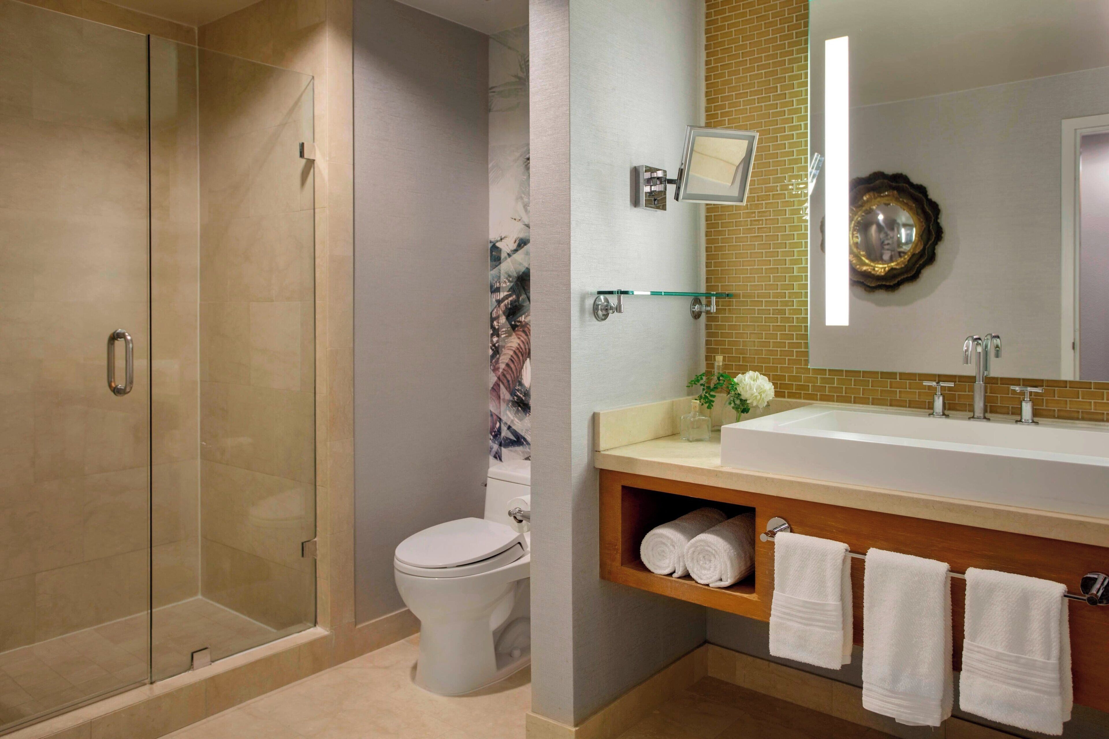A modern bathroom with a glass-enclosed shower, a white toilet, a large sink with a mirror, and neatly rolled towels on a shelf.