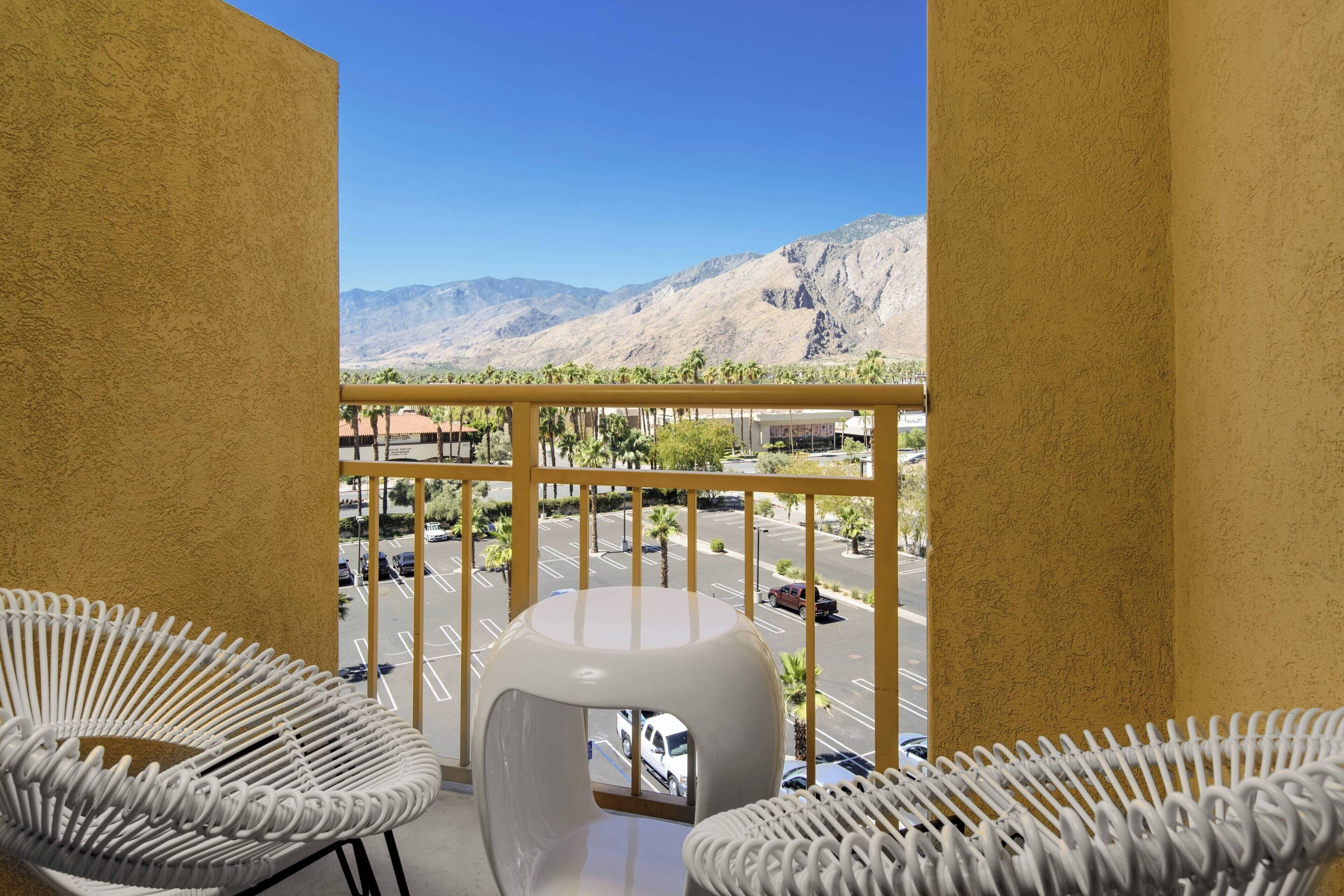 A balcony with yellow walls, white wicker chairs, and a view of a parking lot and mountains.