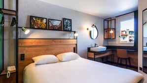 A hotel room with a large bed, a desk with a sink, a mirror, and artwork on the wall. - ibis Pau Centre (Pau)