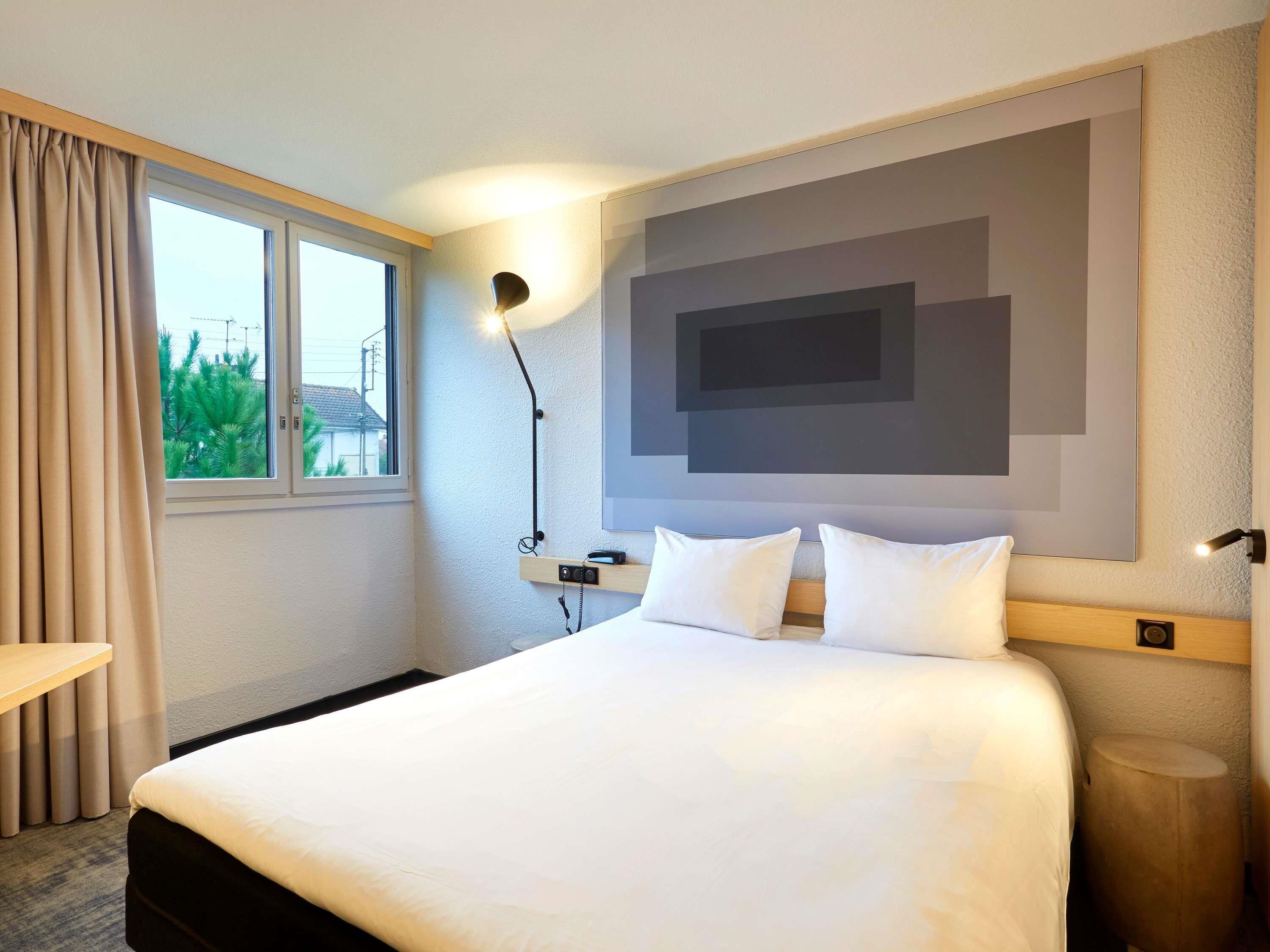 Une chambre d’hôtel comprenant un lit, une table de chevet, une lampe, un téléviseur fixé au mur et une fenêtre avec des rideaux.