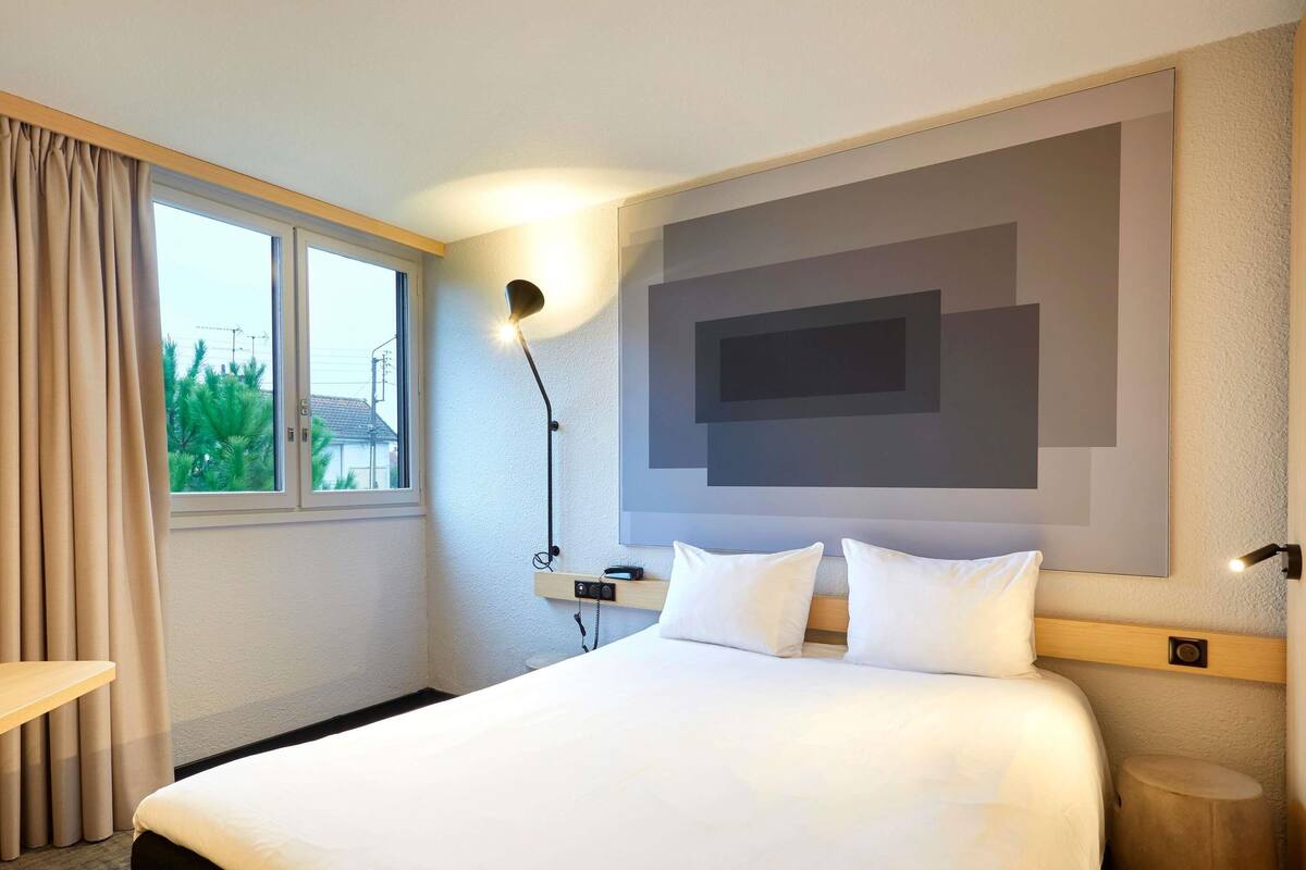 A hotel room with a bed, a bedside table, a lamp, a wall-mounted TV, and a window with curtains.
