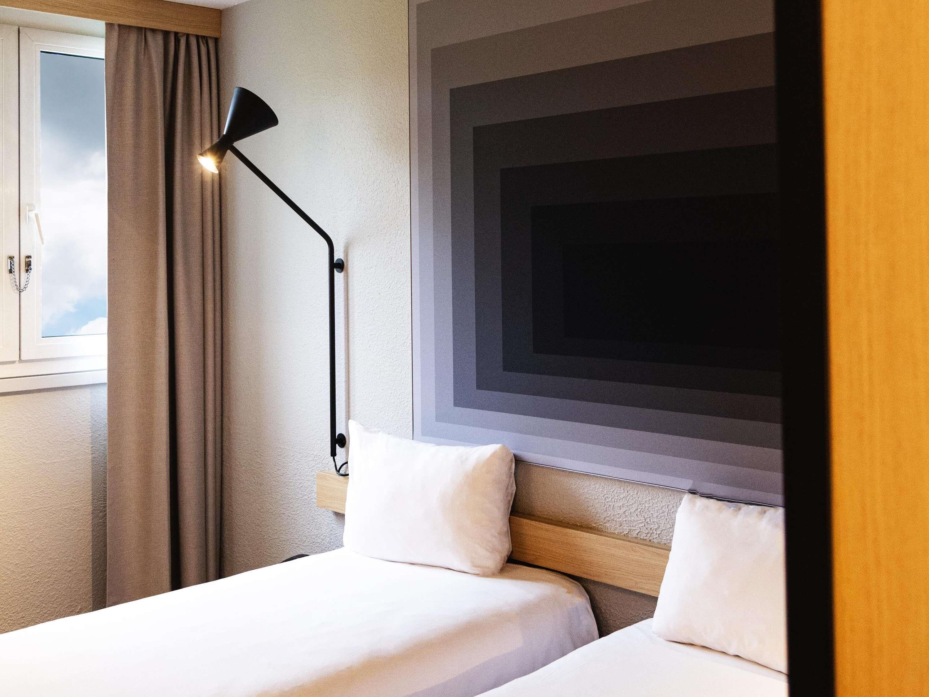 Una habitación de hotel con dos camas, un ventanal con cortinas, una lámpara de pie negra y una pared con diseño geométrico.