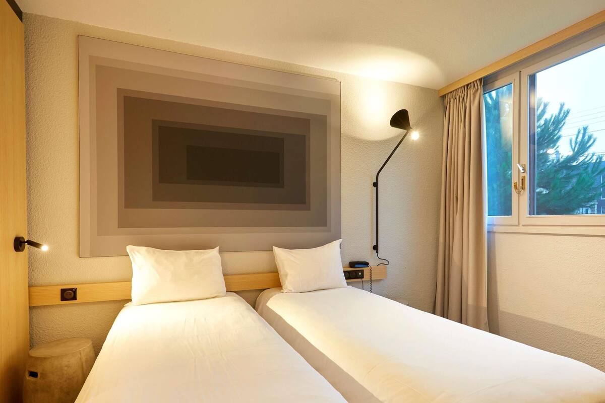 A hotel room with two beds, a large framed artwork above the beds, a bedside lamp, a small bedside table with a phone, and a window with curtains.