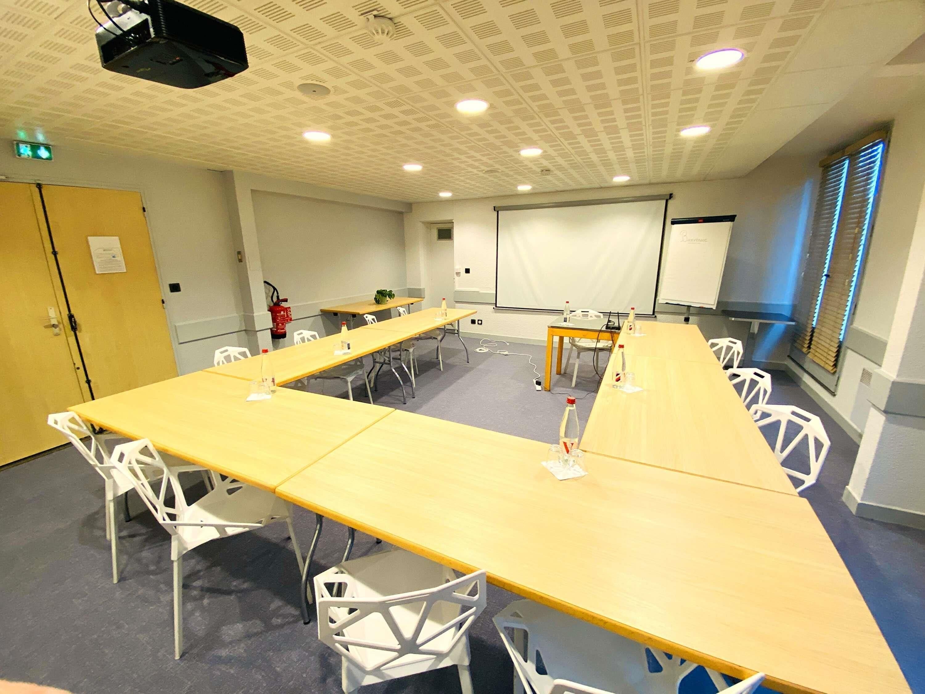 Uma sala de conferências com mesas compridas, cadeiras brancas, um projetor, um quadro branco e um extintor de incêndio.