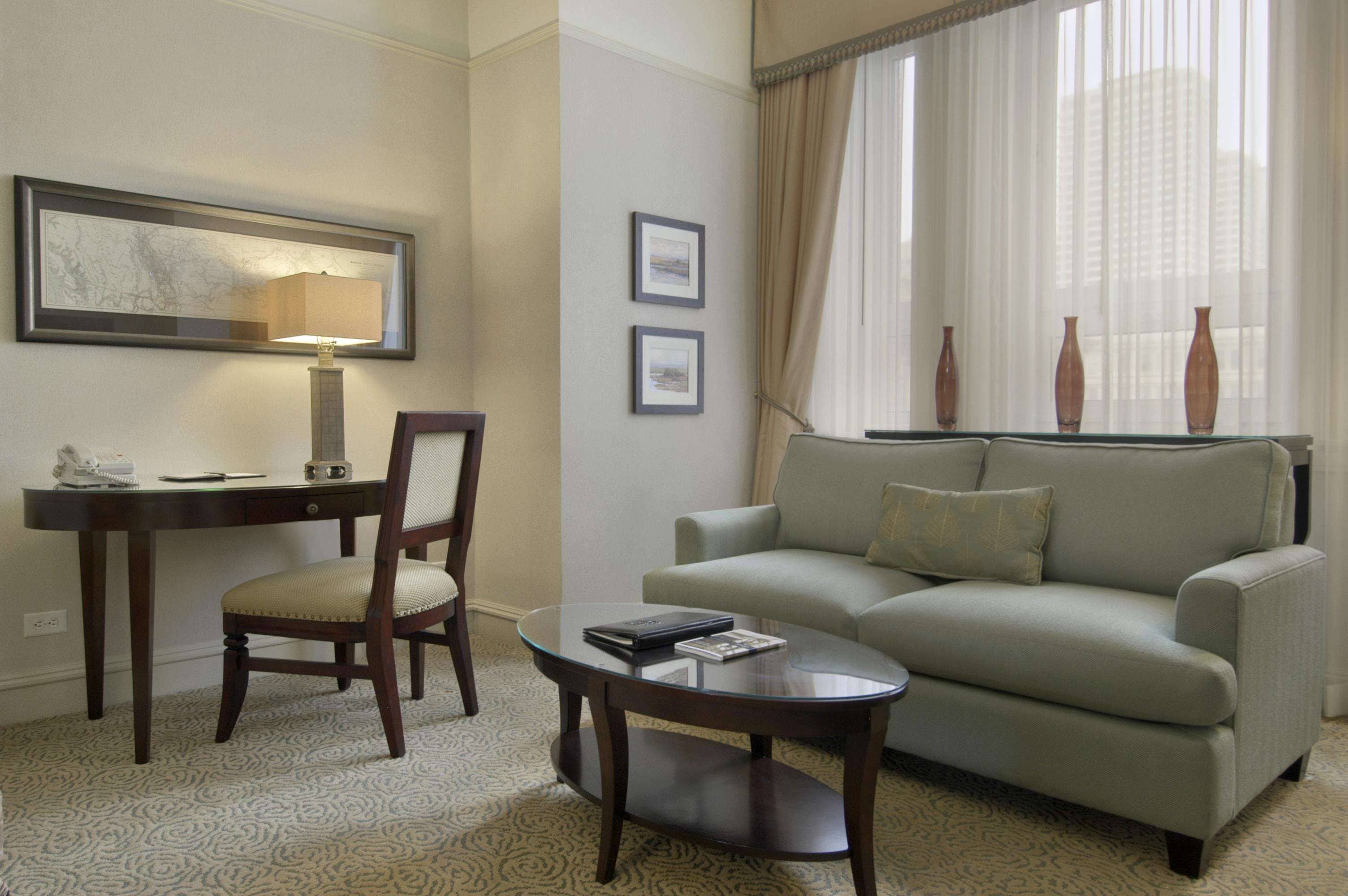 Quarto de hotel com escrivaninha, cadeira, sofá e mesa de centro de vidro.