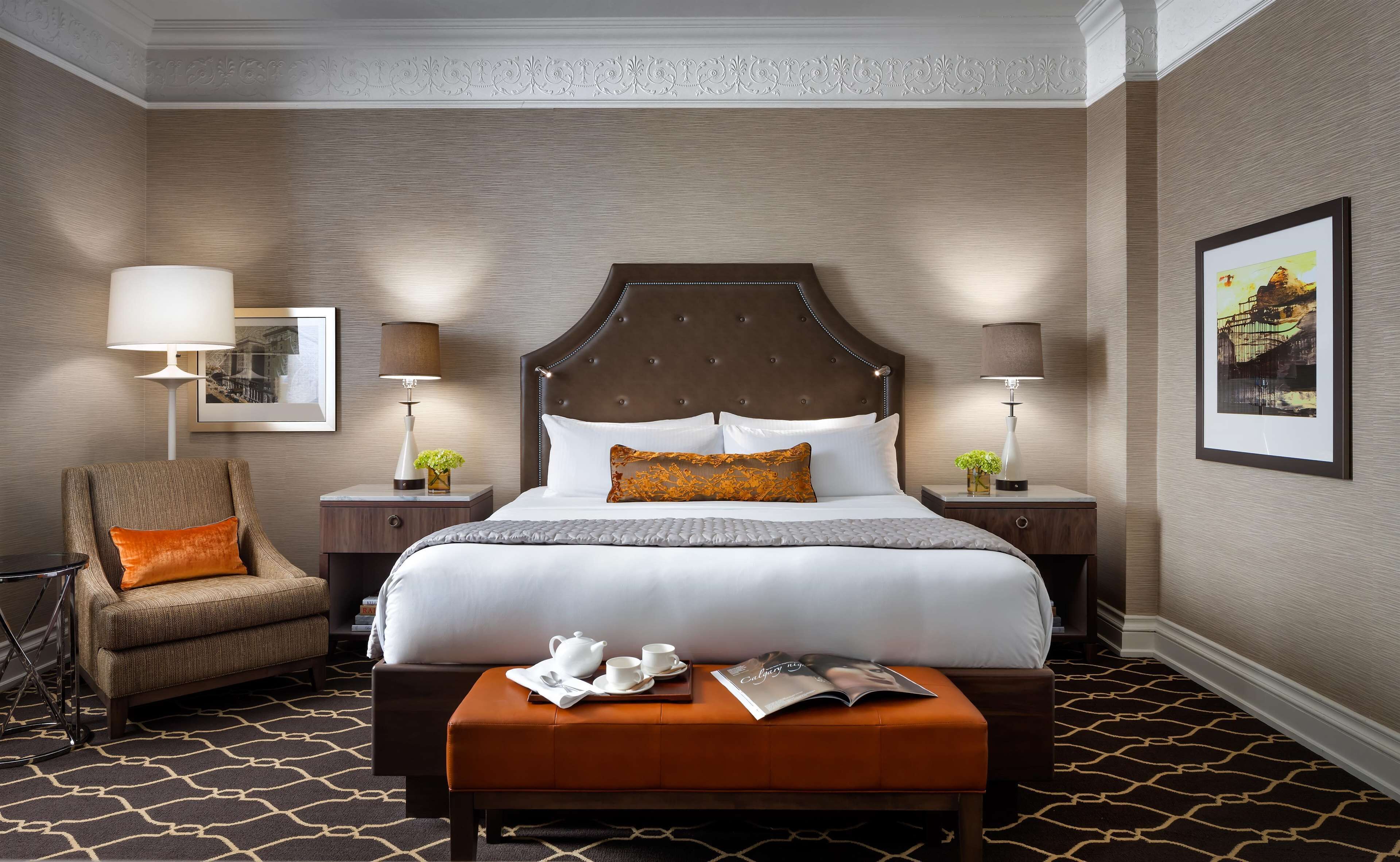 A hotel room with a large bed, two bedside tables with lamps, a chair, a small table with a magazine, and a patterned carpet.