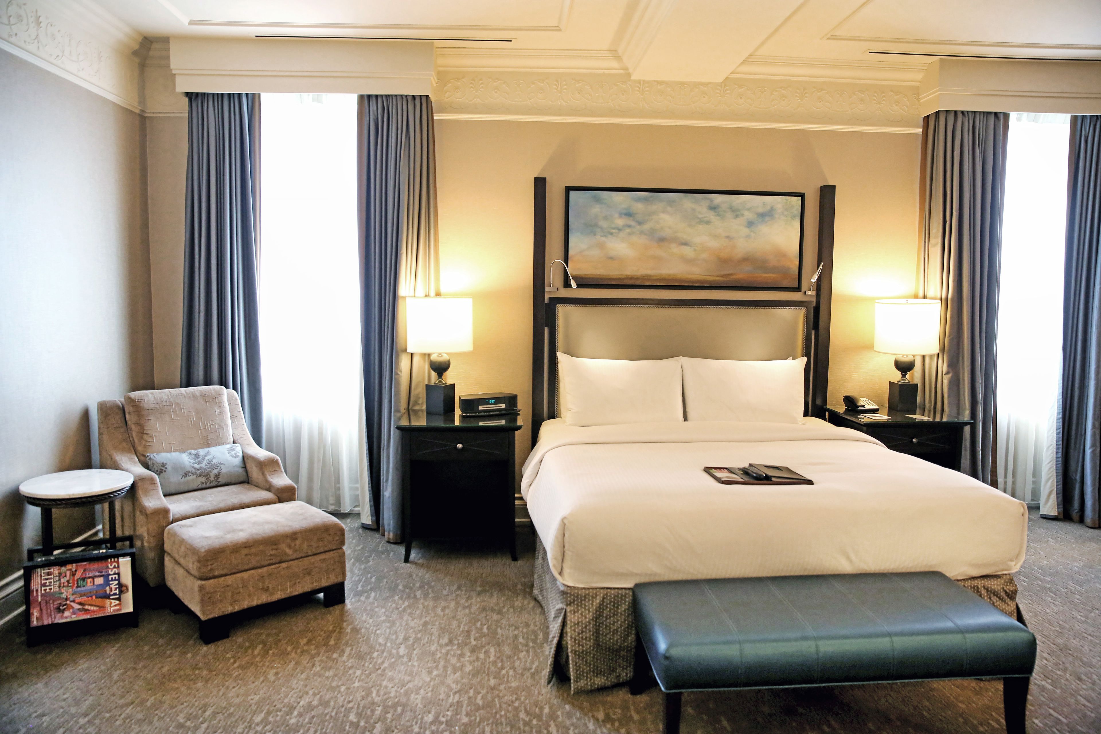 A hotel room with a large bed, a chair, a small table, and a bench.