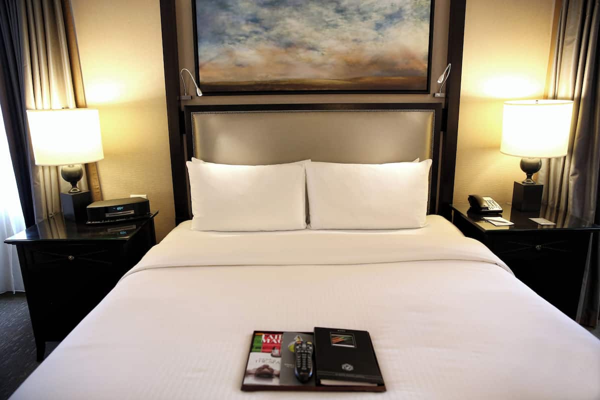 A hotel room with a large bed, two bedside tables, lamps, a painting above the bed, and a book and remote control on the bed.