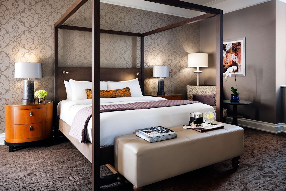 A four-poster bed with a canopy, two bedside tables with lamps, a small round table with a vase and a tray with glasses, and a framed picture on the wall.