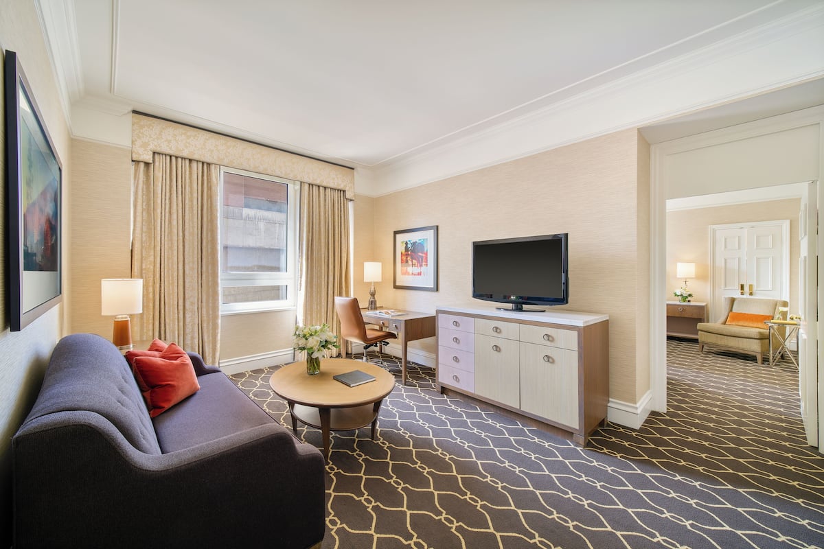 A hotel room with a sofa, a small round table with a vase, a television, a desk, and a cabinet.