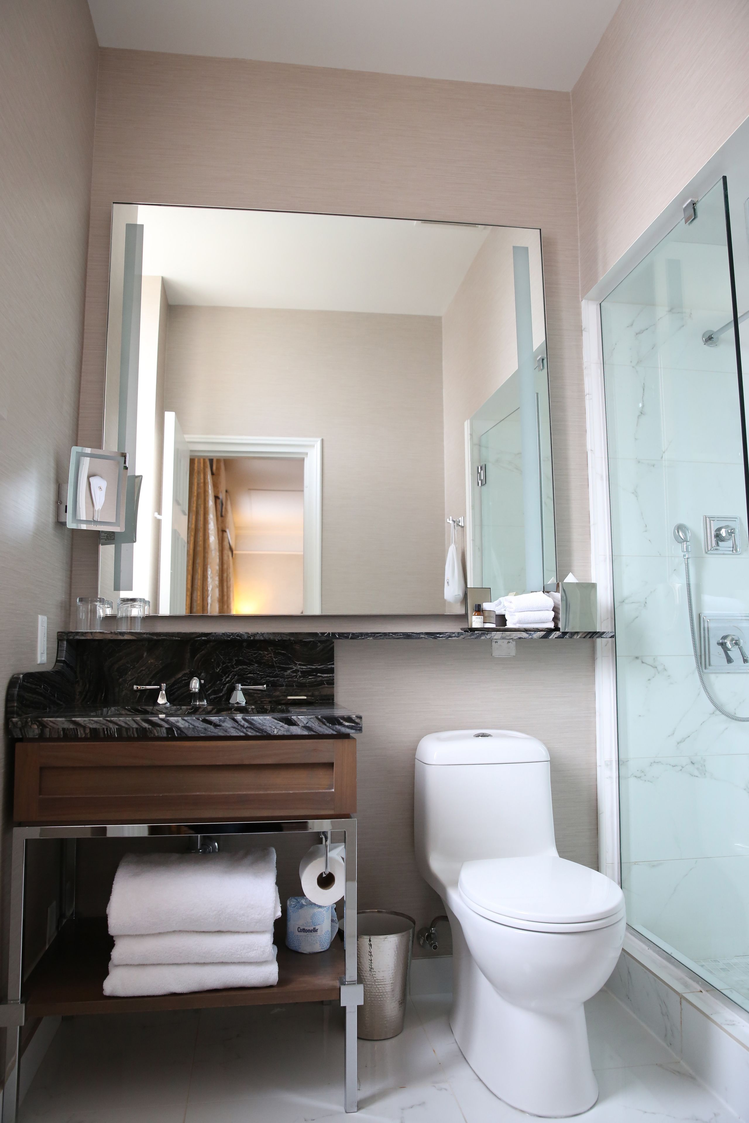Une salle de bain moderne avec un plan de toilette en marbre, des toilettes blanches, une cabine de douche en verre et un grand miroir au-dessus du lavabo.