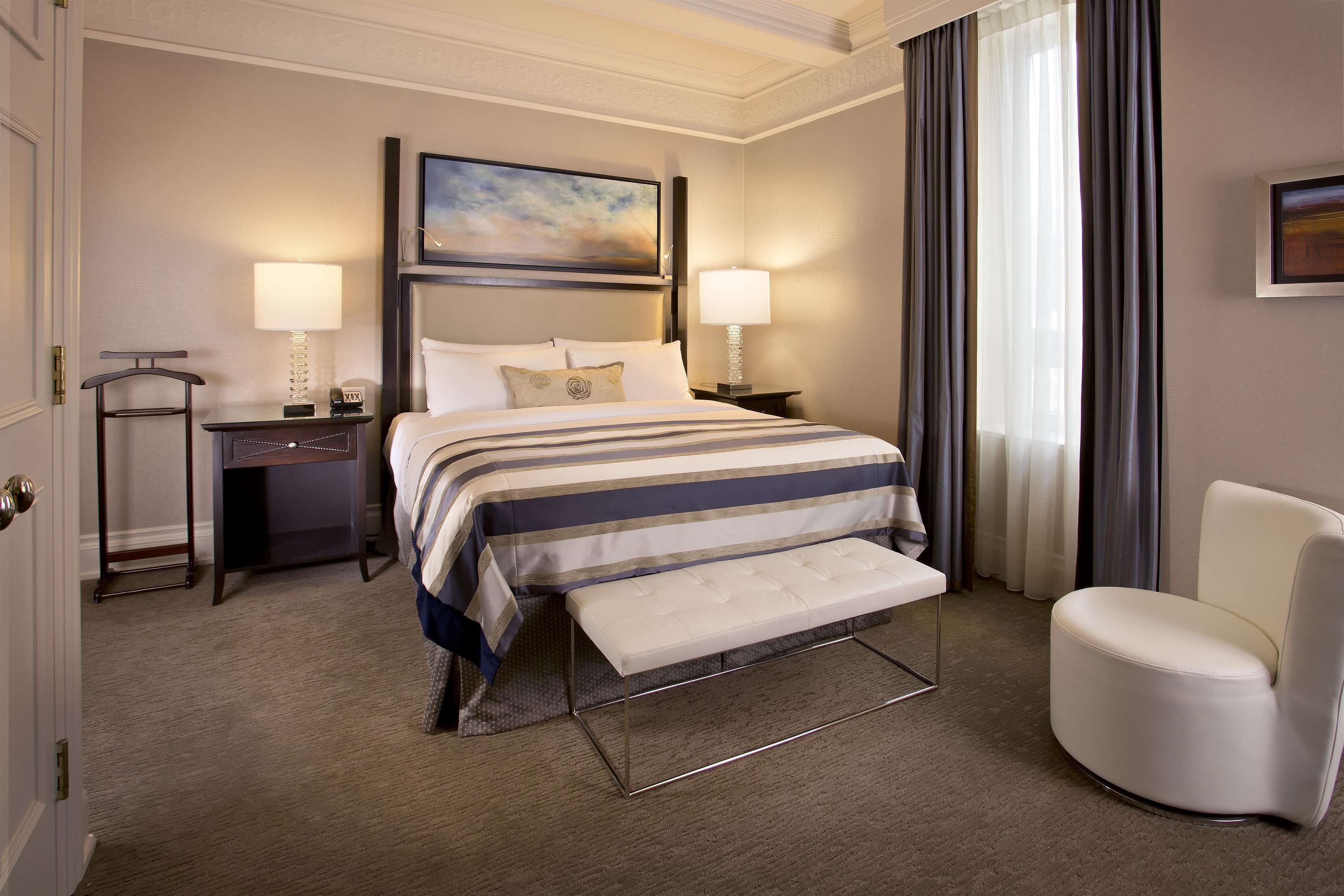 A hotel room with a bed, bedside lamps, a bench, a chair, and a painting on the wall.