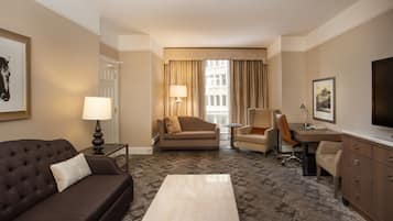A hotel room with a brown sofa, a small table, a desk with a chair, a TV, and a window with curtains.