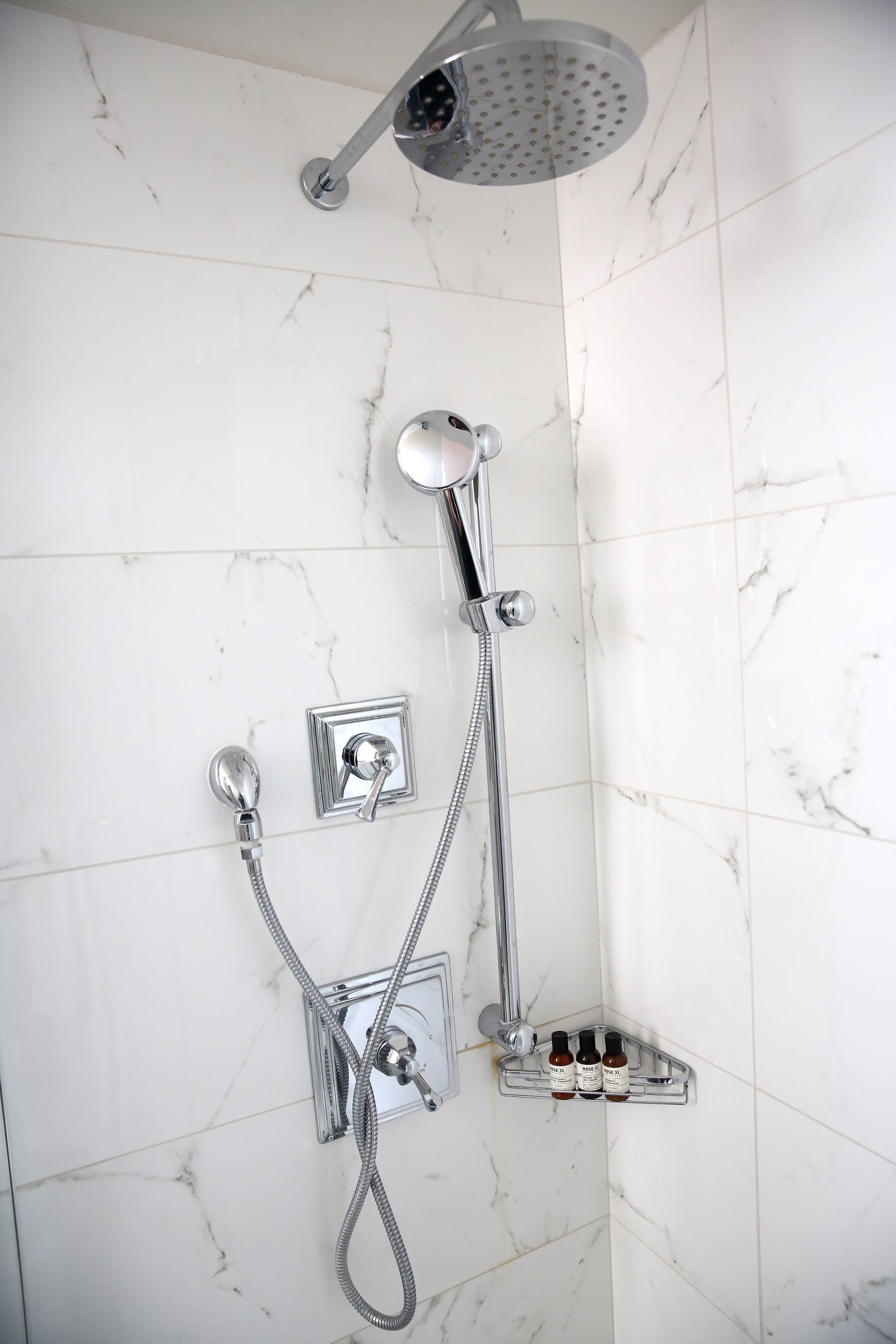 Ducha con cabezal de ducha de mano y grifo empotrado. Hay tres botellas en un estante debajo de la ducha.