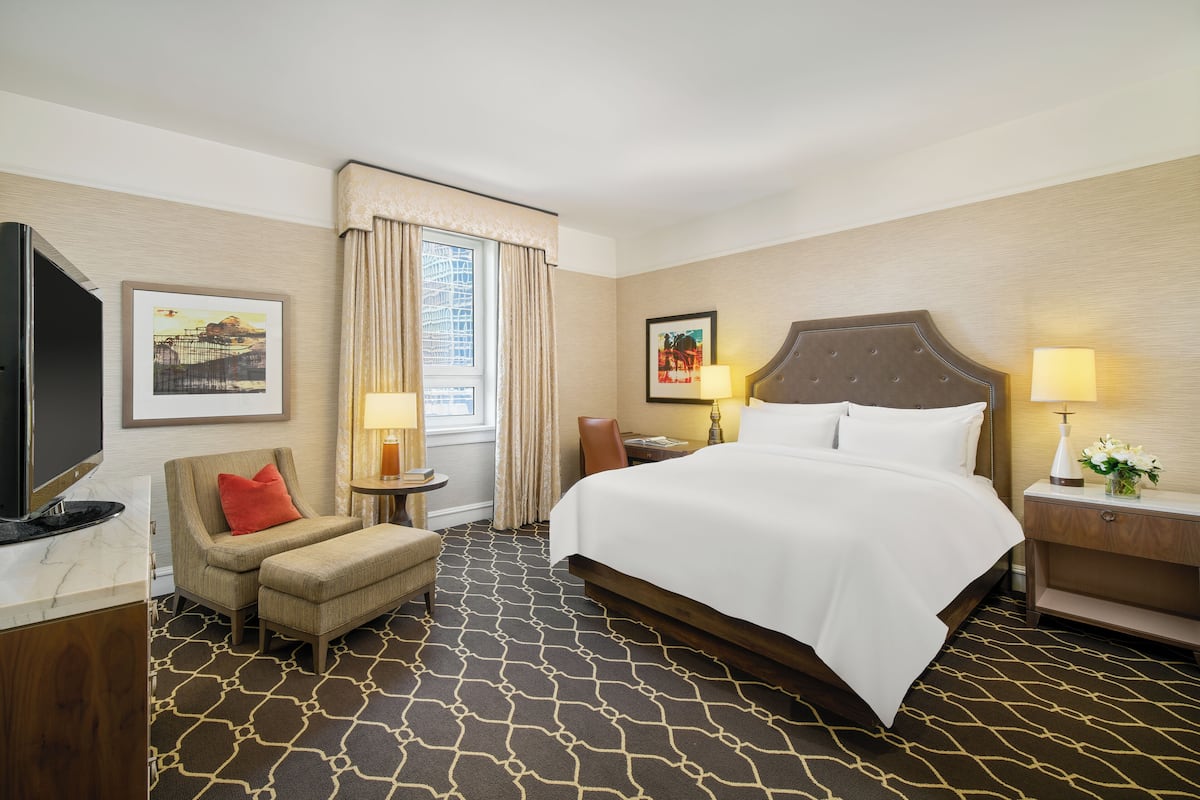 A hotel room with a large bed, a TV, a chair, a desk, and a window with curtains.