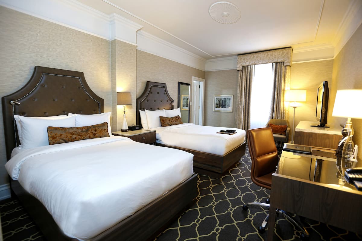 A hotel room with two beds, a desk, a chair, and a TV.
