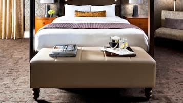 A four-poster bed with a headboard, two bedside tables with lamps, a bedside table with a tray of toiletries, and a footstool.