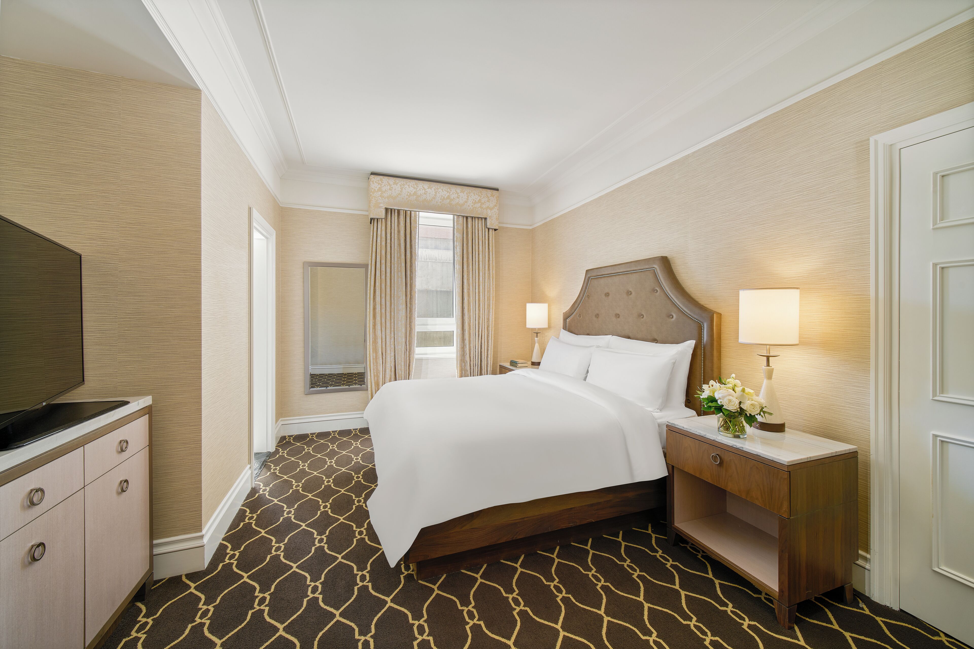 A hotel room with a large bed, bedside lamps, a TV, and a wooden nightstand.