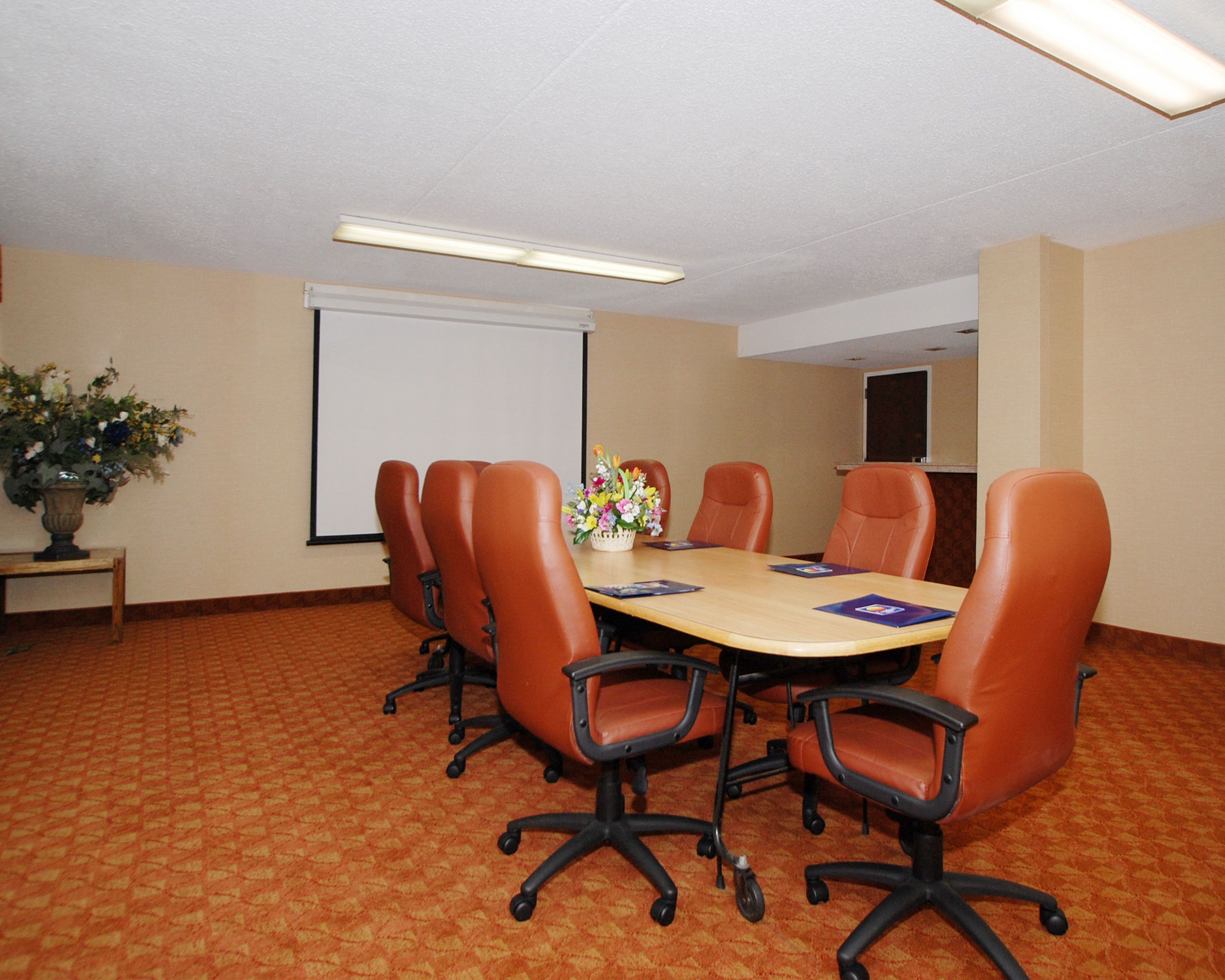 A conference room with a large table, six chairs, a projector screen, and a floral arrangement.