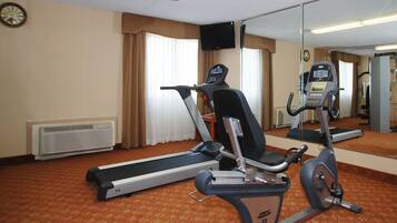 Una habitación de hotel con una cinta de correr, una bicicleta estática, un televisor montado en la pared y un reloj.