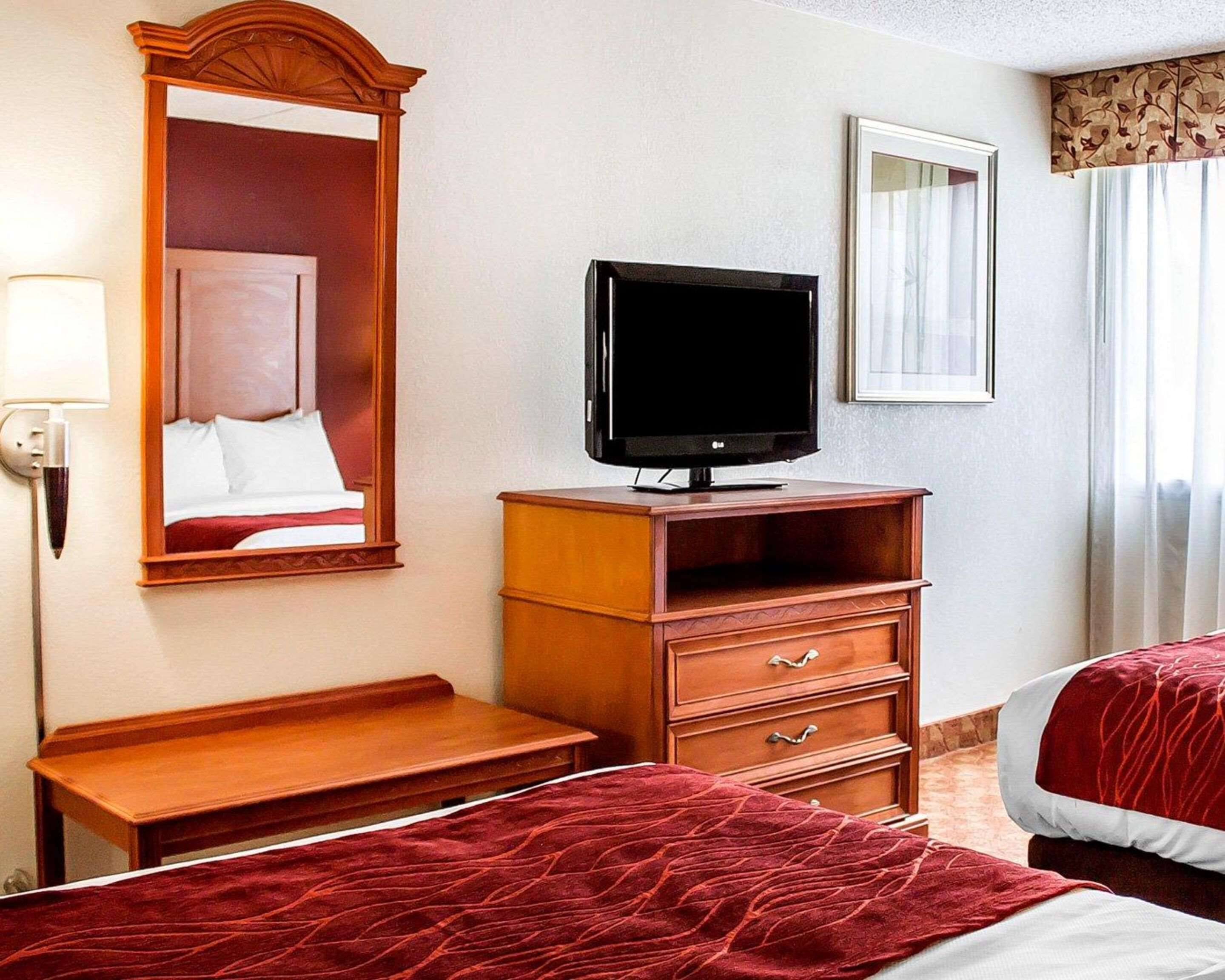 A hotel room with a bed, a TV on a wooden stand, a mirror, and a window with curtains.
