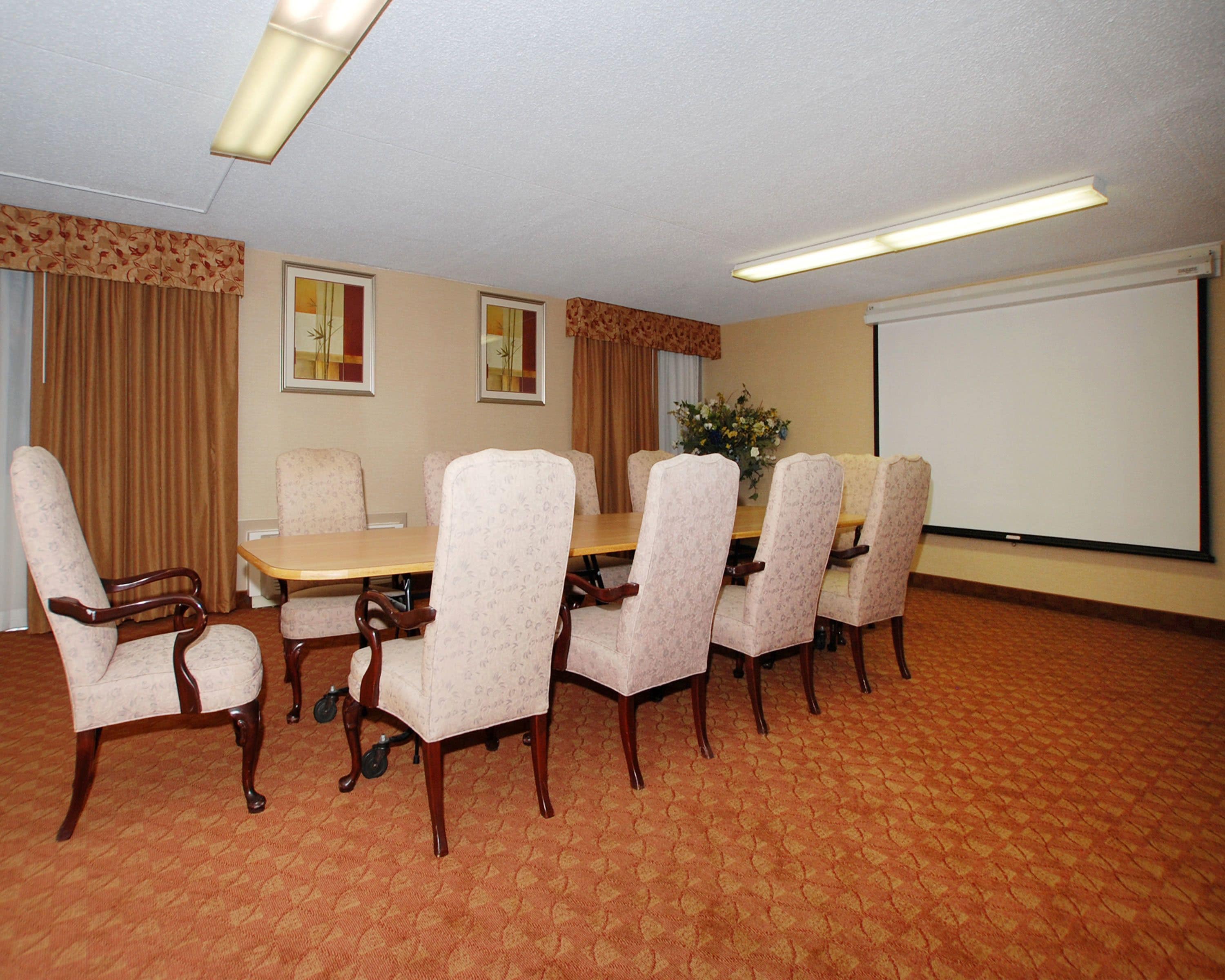 A conference room with a rectangular table surrounded by six upholstered chairs, a projector screen, and a floral arrangement on the table.