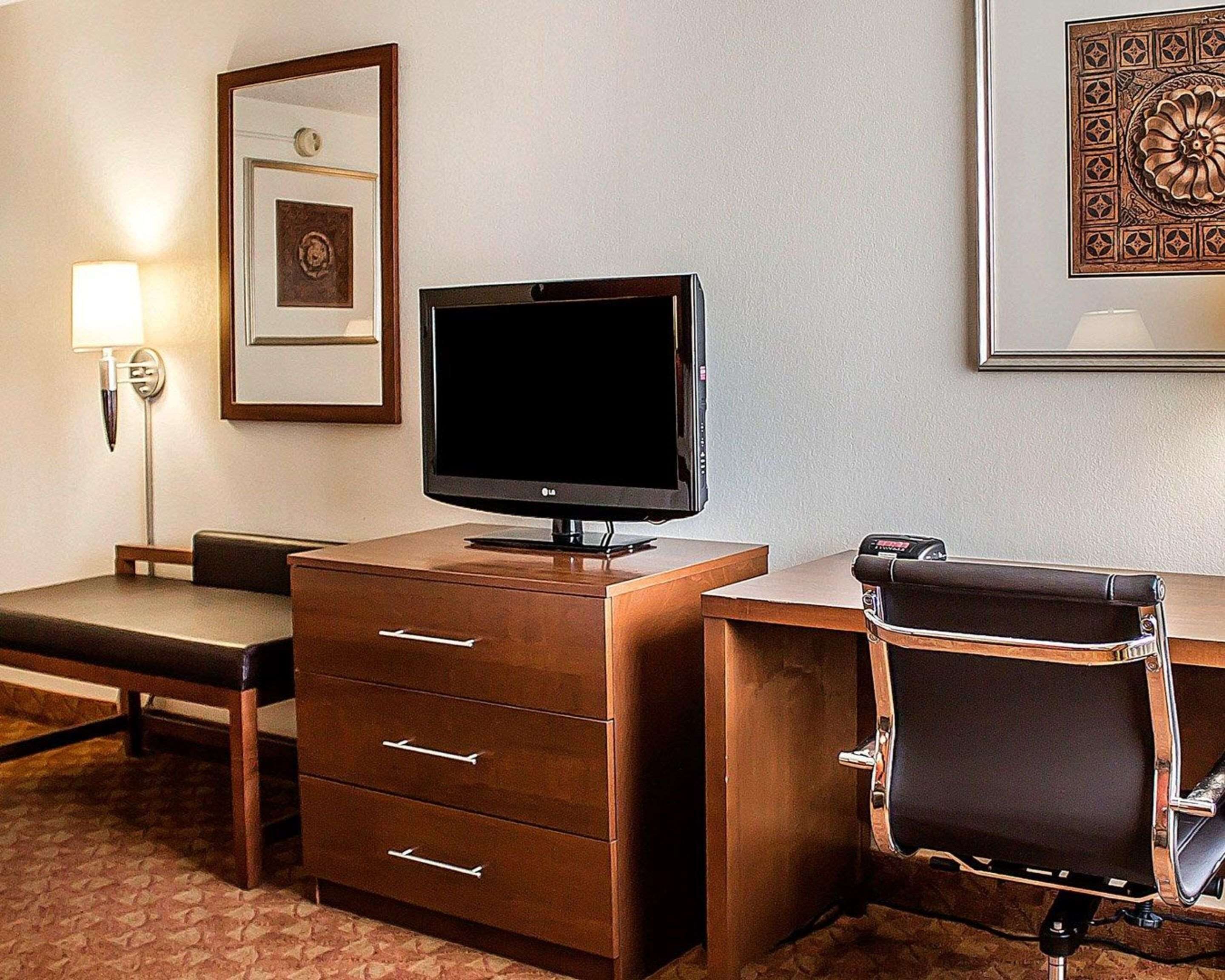 A hotel room with a flat-screen TV on a wooden desk, a leather chair, a desk lamp, and a framed picture on the wall.