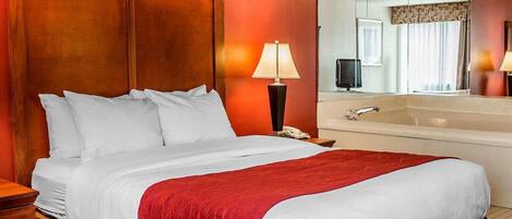 A hotel room with a wooden headboard, a large bed with white bedding and a red throw, a bedside table, a lamp, a mirror, and a bathroom with a bathtub and sink.