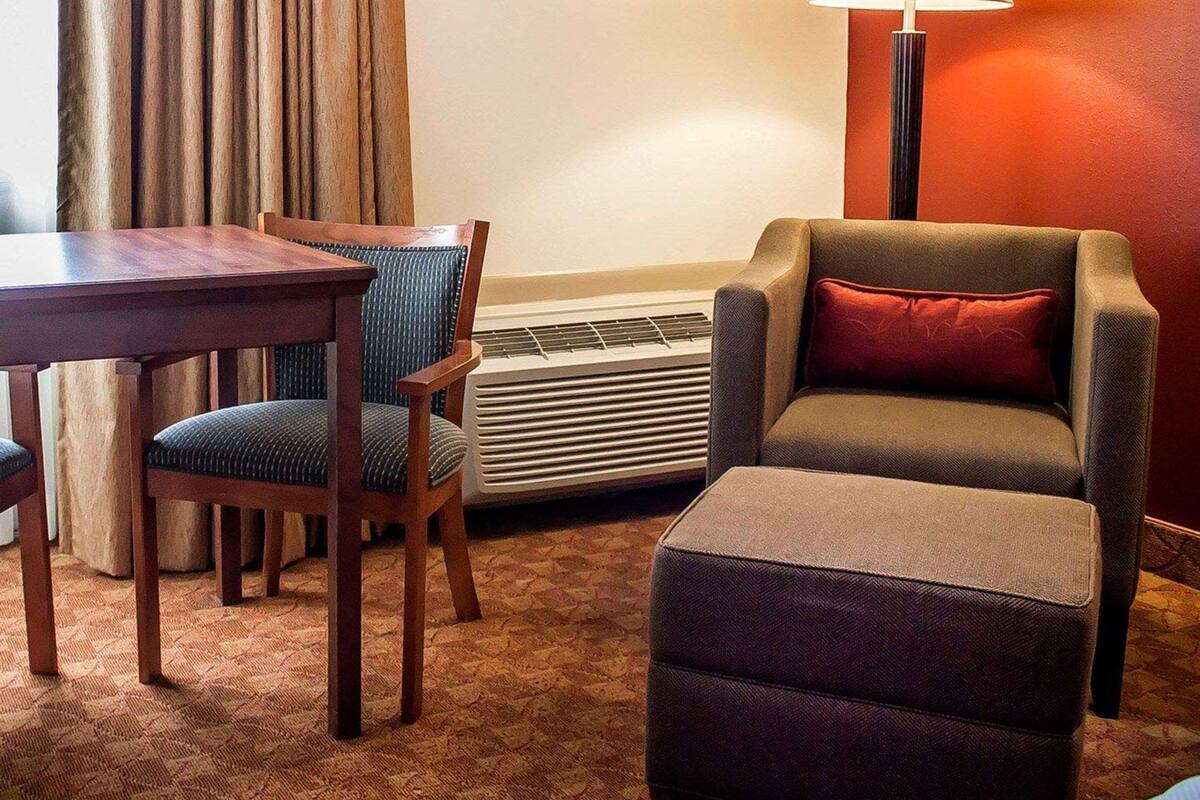 Una habitación de hotel con una mesa de comedor de madera, dos sillas tapizadas, un sofá con un cojín rojo, una lámpara de pie y un calentador fijado a la pared.
