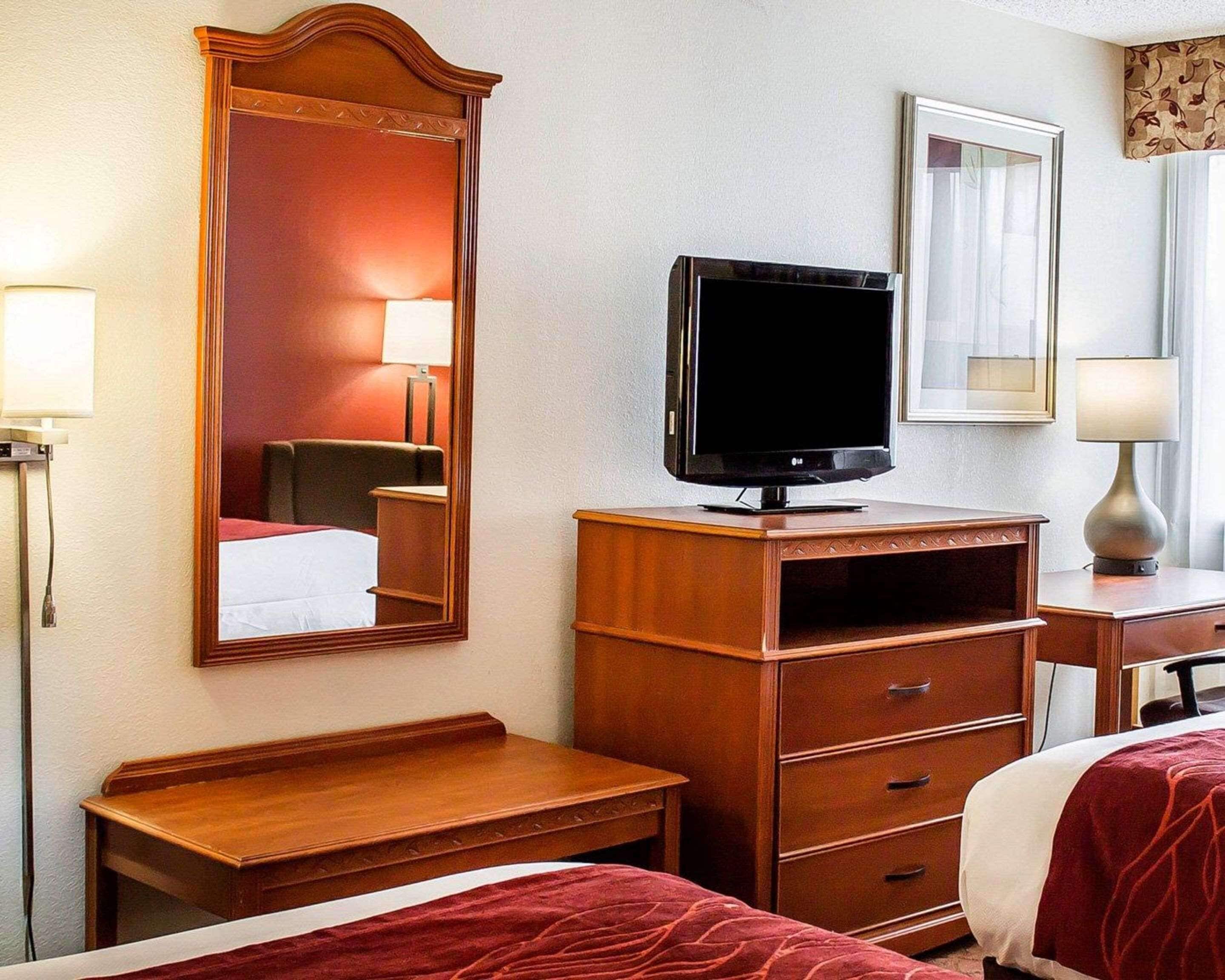 Ein Hotelzimmer mit einem Bett, einem Holztisch, einem Fernseher auf einem Standfuß, einem Spiegel und einem Fenster mit Vorhängen.